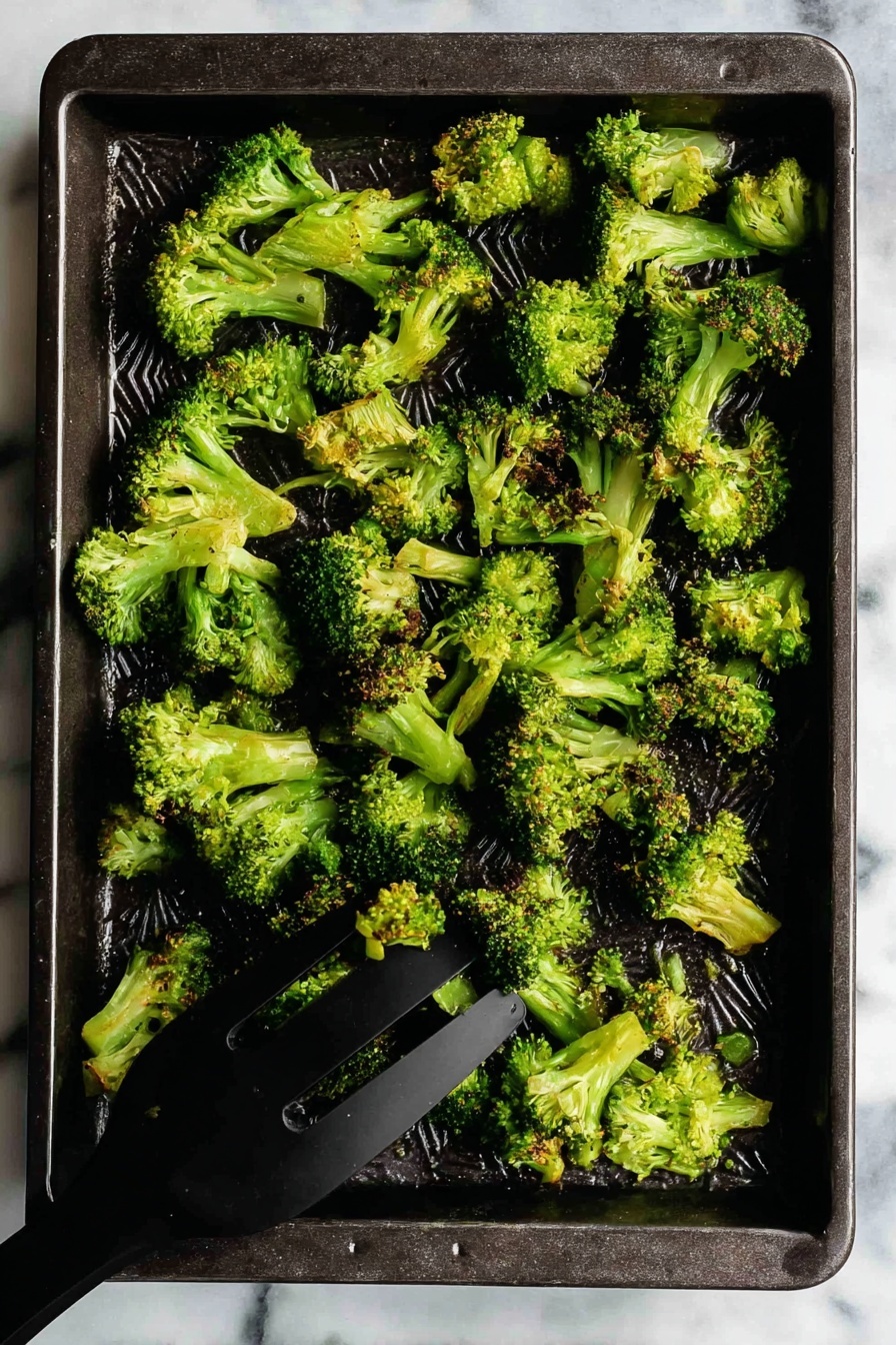 Oven Roasted Broccoli with Garlic and Paprika, roasted broccoli side dish, healthy roasted broccoli, easy vegetable side, flavorful broccoli recipe - A dark rectangular baking tray holds a single layer of cooked broccoli florets. The broccoli pieces are bright green with some slightly browned tips, showing a crispy texture. The florets vary in size and shape, spread unevenly across the tray. In the bottom right corner, a black spatula is lifting some broccoli. The tray rests on a white marbled surface. photo taken with an iphone --ar 2:3 --v 7