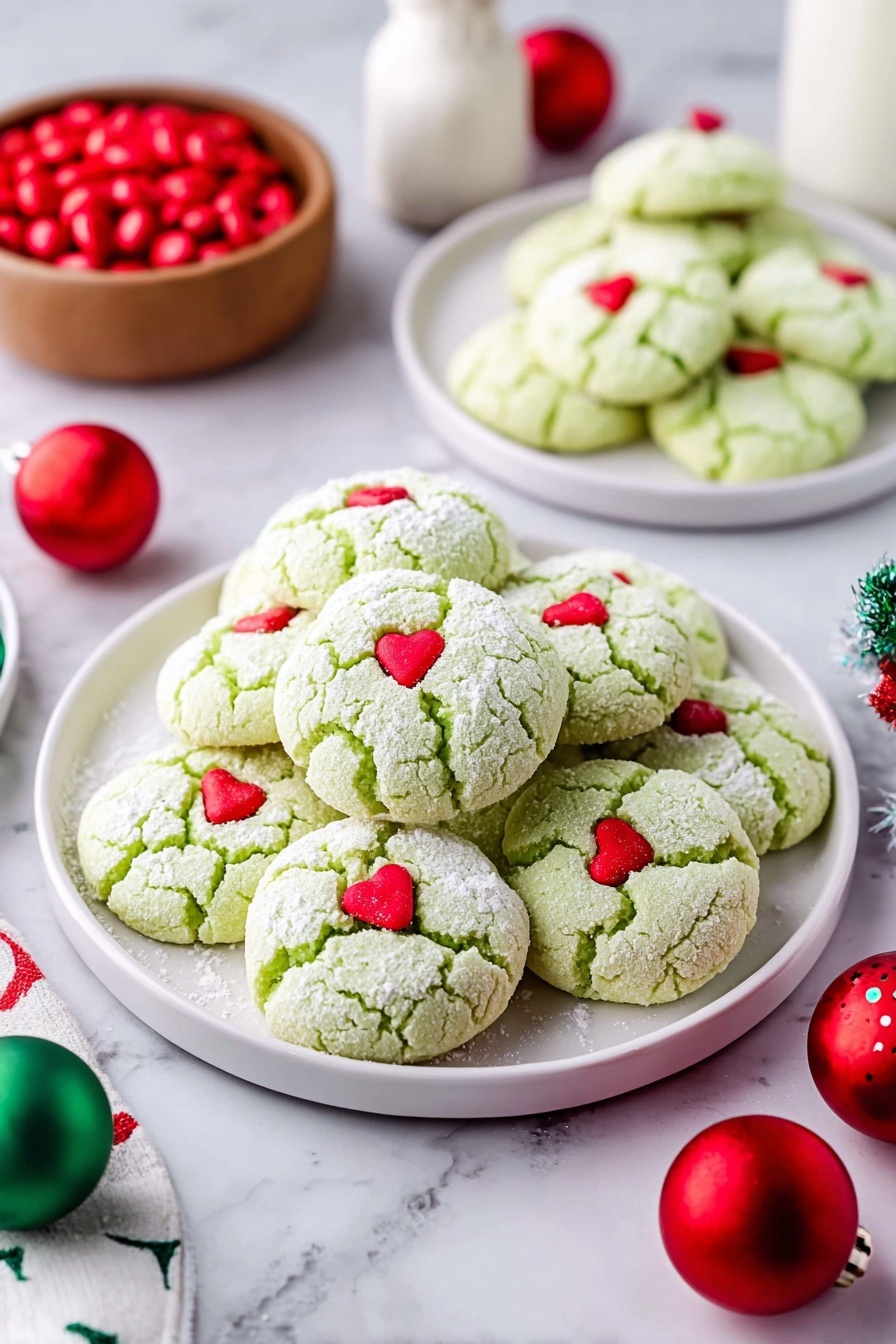 Green Peppermint Cake Mix Cookies, peppermint cookie recipe, festive holiday cookies, easy mint chocolate cookies, winter dessert ideas - A large pile of light green cookies with a soft cracked texture, each topped with a small red heart candy, sits on a white plate in the center. Two smaller white plates in the background hold more of the same cookies. The cookies are dusted lightly with powdered sugar. Around the plates are red and green round Christmas ornaments and a small wooden bowl filled with red candies on a white marbled surface. A white bottle is visible in the background. Photo taken with an iphone --ar 2:3 --v 7
