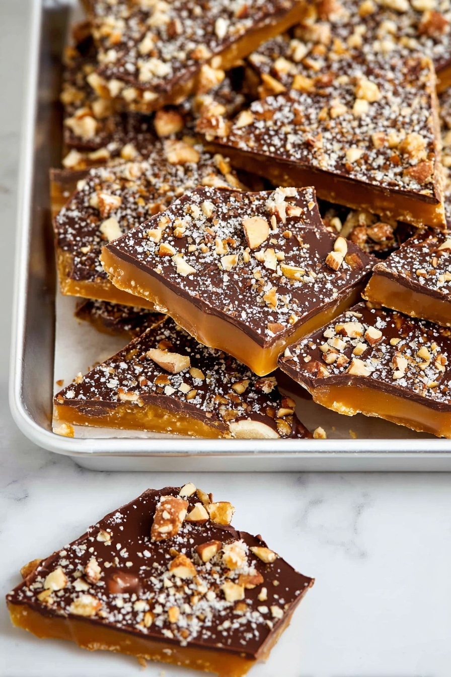 Homemade Toffee with Chocolate and Pecans, chocolate pecan toffee, easy homemade toffee, caramel toffee with nuts, rich chocolate toffee bites - The image shows a white tray full of irregular triangular pieces of candy with three visible layers: a bottom layer of golden caramel, a shiny middle layer of smooth dark chocolate, and a top layer sprinkled heavily with crushed nuts in light brown and white tones. Next to the tray are two more pieces of the same candy, one fully visible and one partly shown, lying on a white marbled surface. The overall look is rich and textured with the nut pieces creating a rough surface on top of the smooth chocolate layer photo taken with an iphone --ar 2:3 --v 7