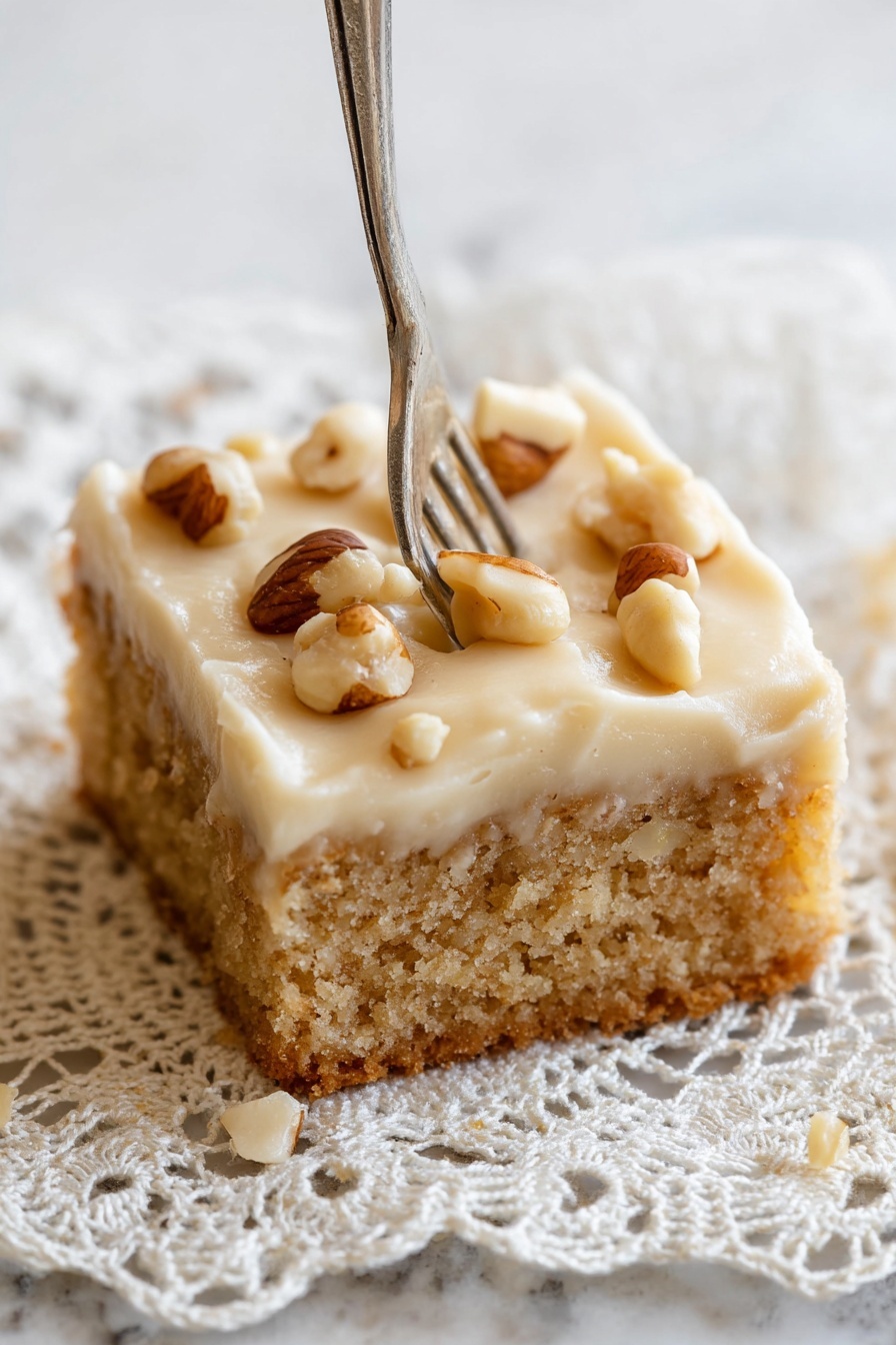 Peanut Butter Cake with Creamy Frosting, peanut butter cake recipe, easy peanut butter cake, moist peanut butter cake, homemade peanut butter cake - A close-up of a single square piece of cake with two layers, the bottom layer is a light brown, soft, crumbly cake base, and the top layer is a thick, pale cream frosting with a smooth texture. The frosting is decorated with whole, shiny brown and white nuts scattered unevenly on top. A fork is pressing slightly into the right side of the frosting. The cake rests on a delicate crochet lace doily with intricate patterns, placed on a white marbled surface. Photo taken with an iphone --ar 2:3 --v 7