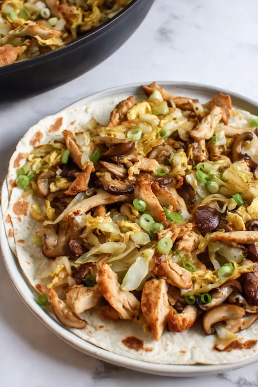 Moo Shu Chicken, Moo Shu Chicken recipe, Chinese chicken stir-fry, quick Asian dinner, easy Chinese chicken dish - This image shows a close-up of a black frying pan filled with a stir-fry dish. The dish has many pieces of brown, cooked chicken strips mixed with light yellow cabbage slices and orange-yellow egg strips. There are also some sliced mushrooms with a dark brown outer edge and light beige inside, scattered throughout. Small green onion pieces are sprinkled over the dish adding a splash of green color. The frying pan is set on a white marbled surface. photo taken with an iphone --ar 2:3 --v 7