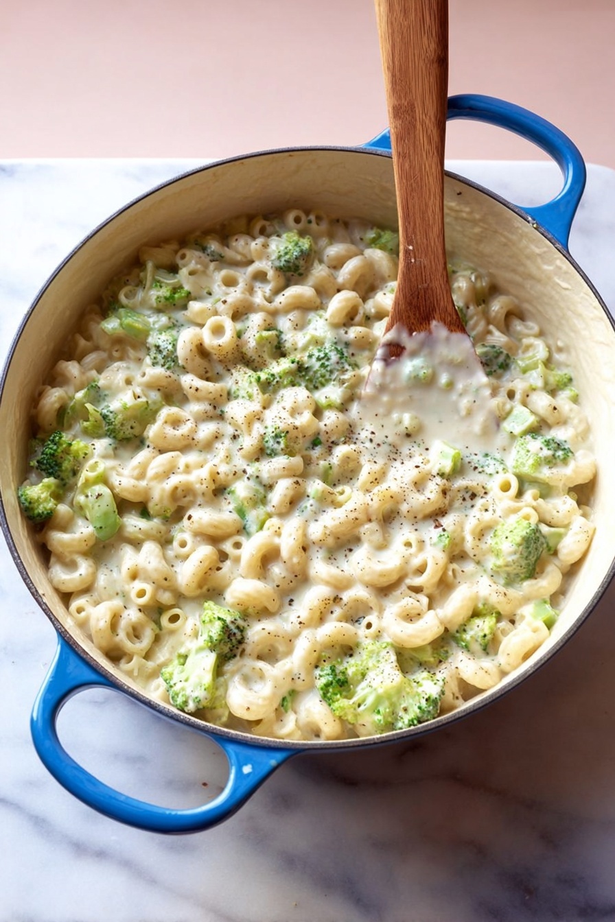 Creamy Broccoli Pasta, creamy broccoli pasta recipe, easy broccoli pasta, cheesy broccoli pasta dish, quick vegetable pasta - A large white pot with blue handles is filled with small tube pasta and small green broccoli pieces mixed in a thick, creamy white sauce with visible black pepper specks, all stirred by a wooden spoon standing upright in the pot, placed on a white marbled surface with a soft pink background, photo taken with an iphone --ar 2:3 --v 7