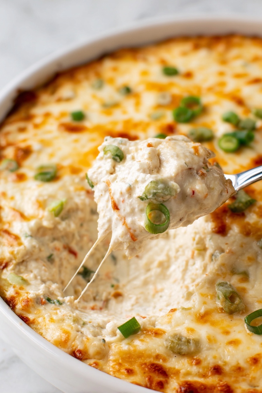Crab Rangoon Dip, Crab Rangoon Dip with Crispy Wontons, cheesy crab dip, savory appetizer dip, crab and cream cheese dip - A close-up of a white dish filled with a creamy dip that has a thick melted cheese layer on top. The cheese is golden with light brown spots and sprinkled with small green onion pieces. The creamy layer underneath is light beige with visible small bits of other ingredients, showing a rich and creamy texture. A spoon is lifting a scoop of the dip, revealing the thick inside. The dish is on a white marbled surface. photo taken with an iphone --ar 2:3 --v 7