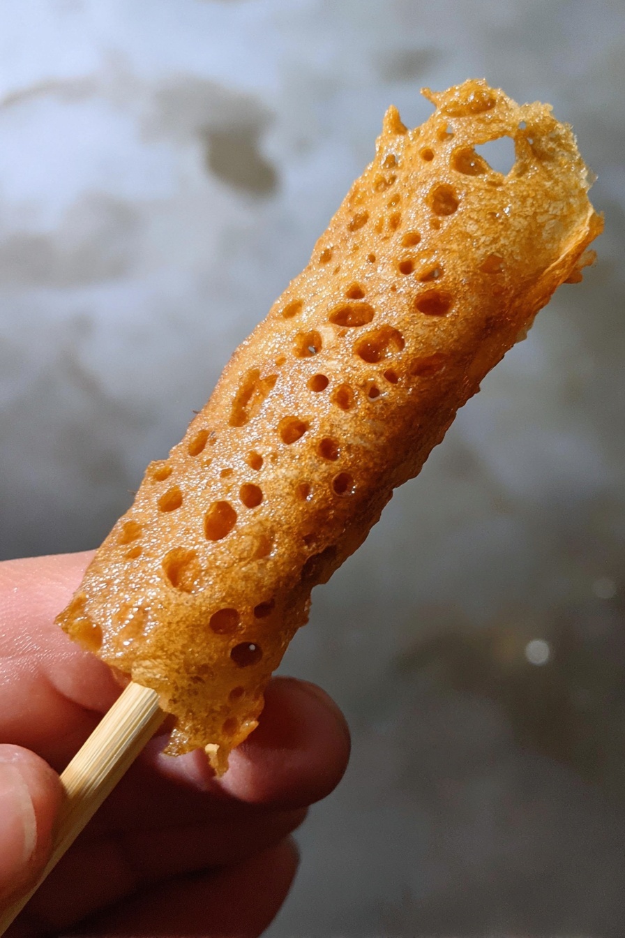 Brandy Snaps, delicate crispy treats, homemade brandy snaps, easy dessert recipes, holiday sweets - A close-up image of a golden brown, thin, lace-like fried snack wrapped around a light wooden stick held by a woman's hand, with a textured, crispy surface that has small holes and a shiny finish, against a background with a white marbled texture. photo taken with an iphone --ar 2:3 --v 7