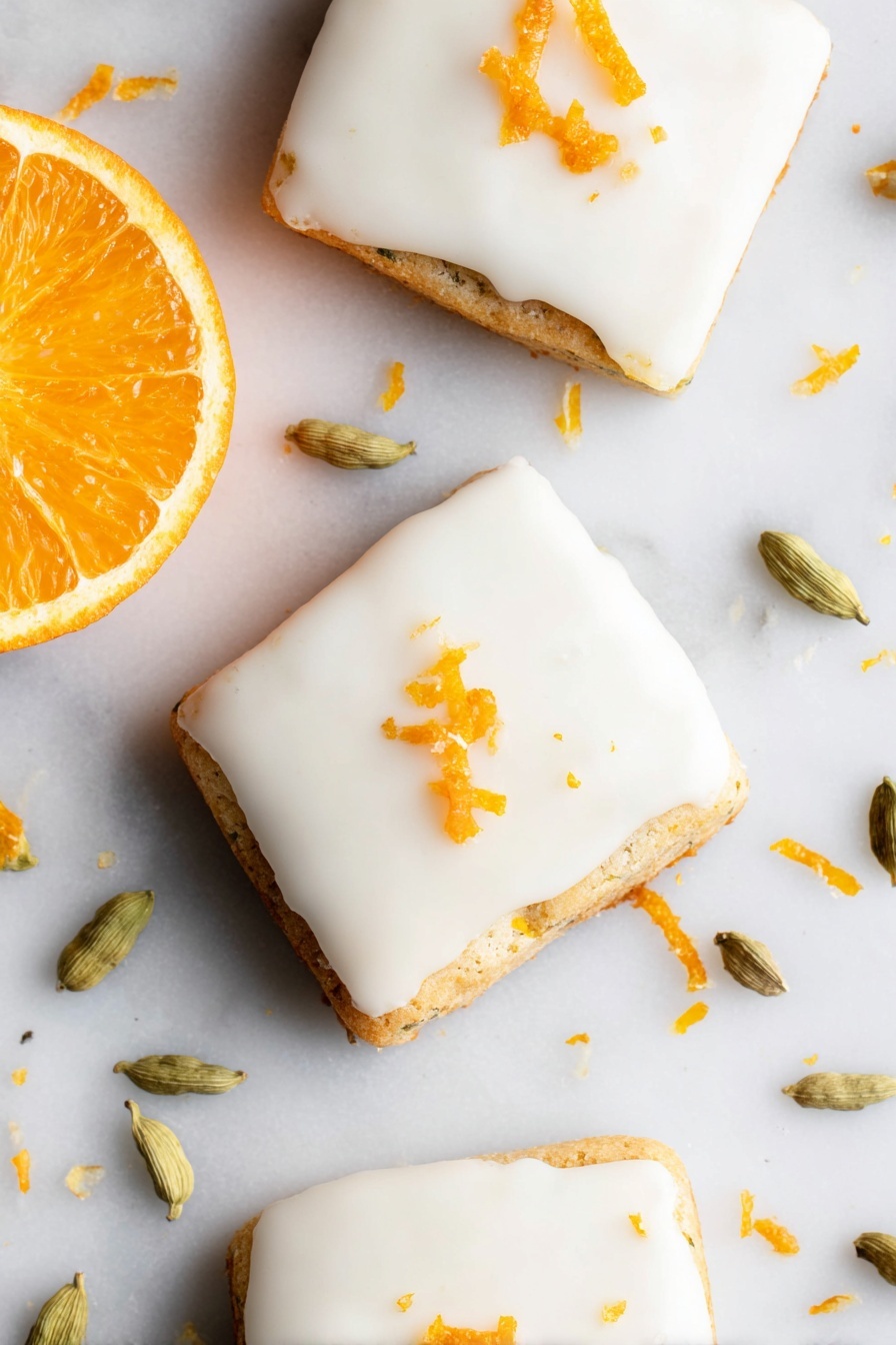 Cardamom Shortbread Cookies with Orange Glaze, fragrant shortbread cookies, orange glaze cookie recipe, aromatic holiday cookies, easy shortbread with citrus glaze - The image shows three square-shaped cookies with smooth white icing covering the top layer. Each cookie is slightly thick, with the base layer visible under the icing, showing a light golden-brown color and a slightly textured edge. Small pieces of orange zest are scattered on top of the icing, adding a bright orange color contrast. To the left side of the cookies, there is a fresh orange half with a vibrant, juicy orange color. Around the cookies on the white marbled surface, there are several light green cardamom pods and scattered bits of orange zest. The photo taken with an iphone --ar 2:3 --v 7
