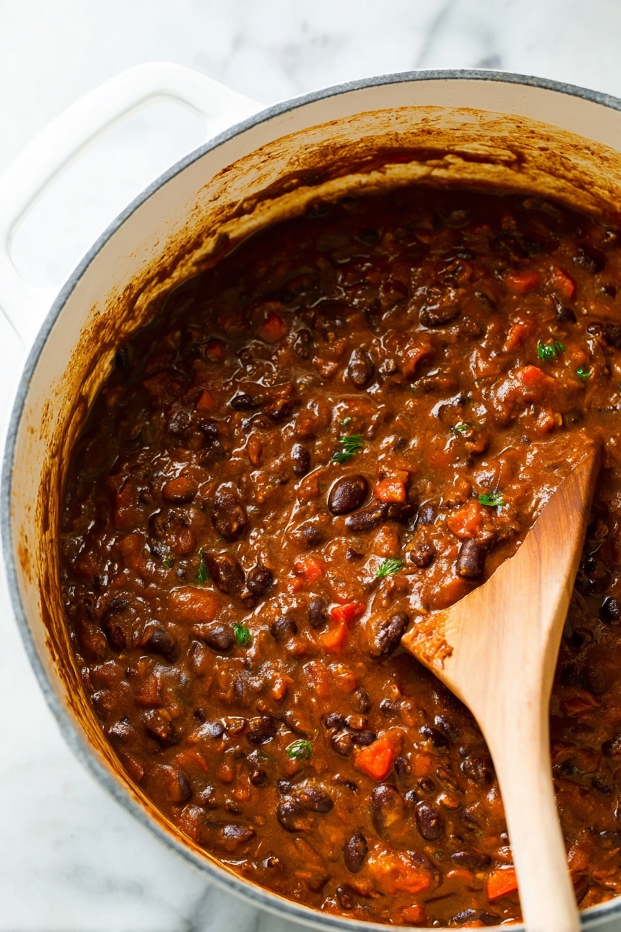 Vegetarian Chili, Best Vegetarian Chili, Hearty Vegetarian Chili, Easy Vegetarian Chili Recipe, Healthy Vegetarian Chili - A close-up view of a thick stew-like dish inside a large white pot with a white marbled surface underneath. The dish has a rich brown color with visible layers of ingredients, including dark brown beans, small red chunks, and some green herb bits mixed throughout. A wooden spoon is partly dipped in the stew on the right side, showing the texture which is soft and slightly chunky with a glossy finish. The sides of the pot have some sauce splashes and the scene looks warm and hearty. Photo taken with an iphone --ar 2:3 --v 7