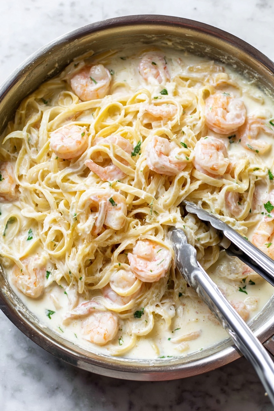 Shrimp Alfredo Pasta, creamy shrimp pasta, easy seafood pasta, quick Shrimp Alfredo, cheesy shrimp pasta - A close-up view of a silver pan filled with creamy white sauce pasta mixed with pink shrimp. The pasta, light yellow and smooth, is covered in a thick white sauce and evenly spread throughout the pan. Plump shrimp are scattered on top and mixed in, their soft pink color contrasting with the white sauce. Small green herb pieces are sprinkled over the dish for a fresh touch. A pair of metal tongs and a spoon are resting on the pasta, indicating the dish is being stirred or served. The pan sits on a white marbled surface. photo taken with an iphone --ar 2:3 --v 7