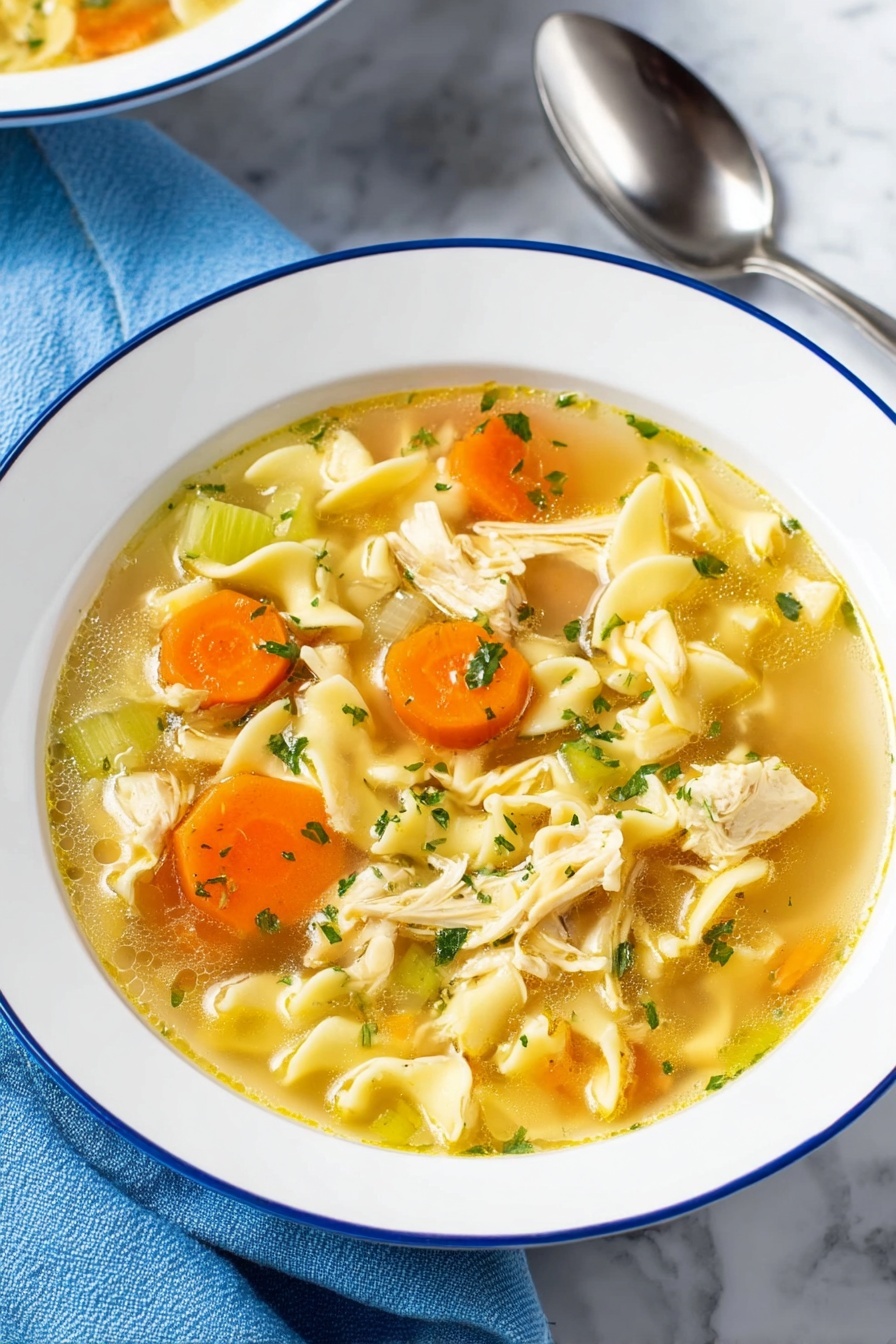 Vegan Chicken Noodle Soup, vegan noodle soup, plant-based chicken soup, vegan comforting soup, dairy-free chicken noodle soup - A bowl of chicken noodle soup is shown with clear, yellowish broth filling the white bowl with a thin blue rim. The soup has several layers including bright orange carrot slices and pale green celery pieces scattered throughout. Soft, wavy egg noodles are curled in different shapes, mixed with small chunks of cooked chicken that are light beige with a tender texture. Small green parsley flakes are sprinkled on top, adding a fresh touch. The bowl sits on a white marbled surface with a blue cloth and shiny silver spoon nearby. photo taken with an iphone --ar 2:3 --v 7