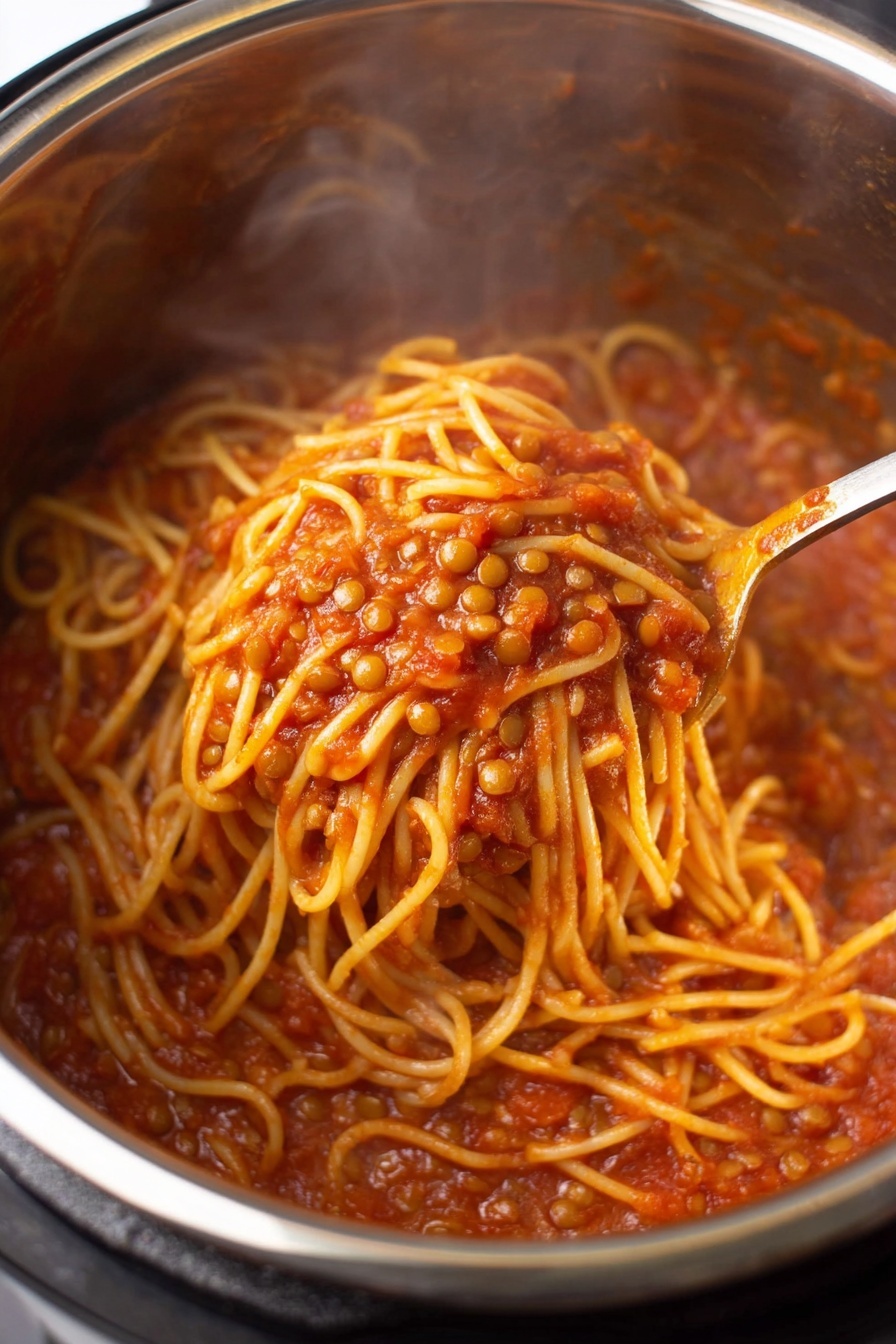 Instant Pot Lentil Spaghetti, healthy lentil pasta, quick vegan dinner, easy Instant Pot recipes, protein-packed pasta dishes - A close-up view inside a shiny stainless steel pot filled with spaghetti noodles mixed with small lentils in a rich red tomato sauce; the noodles have a soft yellow color and are coated well by the thick, slightly chunky sauce; a spoon is stirring the mixture, showing a tangled clump of noodles lifted above the rest with pieces of lentils visible around; steam rises gently from the hot dish. photo taken with an iphone --ar 2:3 --v 7