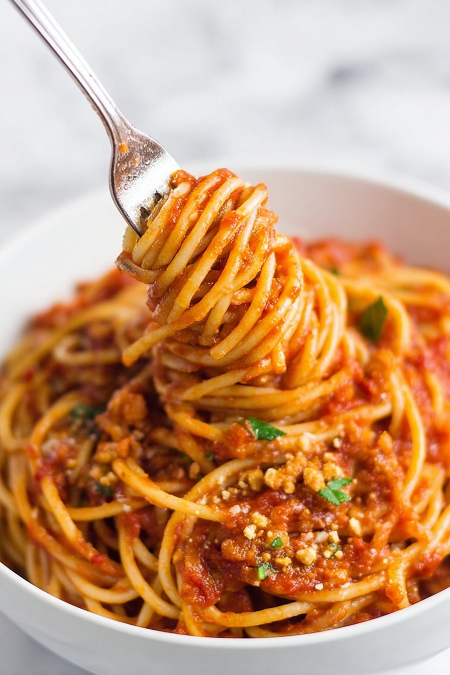 Instant Pot Lentil Spaghetti, healthy lentil pasta, quick vegan dinner, easy Instant Pot recipes, protein-packed pasta dishes - A close-up of a white bowl filled with spaghetti noodles coated in rich red tomato sauce with visible small chunks and texture throughout. The spaghetti is thick and twisted around an inserted fork in the center, lifting the noodles upward, showing a dense mix of sauce and soft pasta strands. Small bits of green herbs and crumbled light brown topping are scattered on the surface, adding color contrast. The bowl is placed on a white marbled surface, and the image has a soft background with a focus on the noodles and sauce. photo taken with an iphone --ar 2:3 --v 7