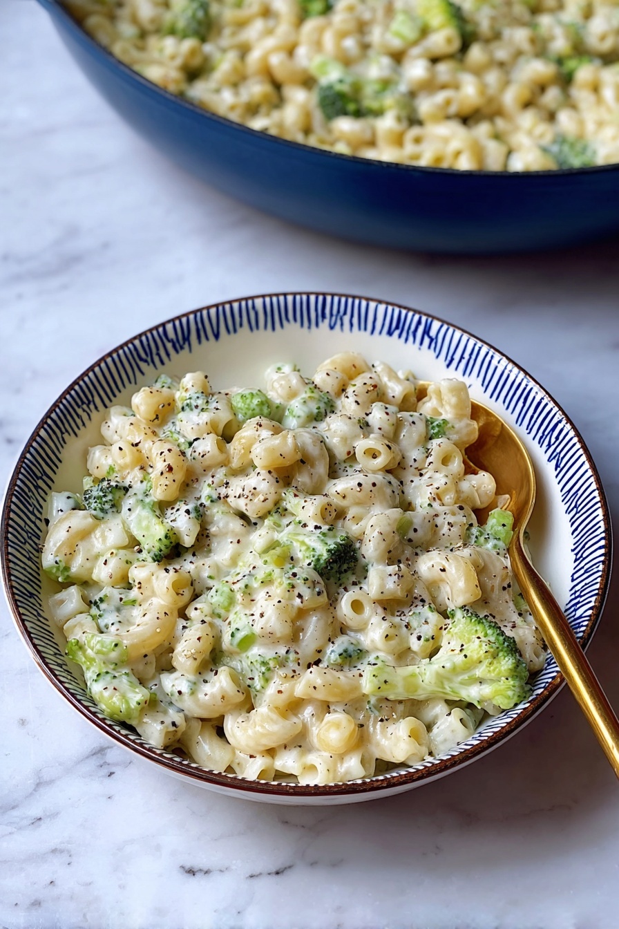 Creamy Broccoli Pasta, creamy broccoli pasta recipe, easy broccoli pasta, cheesy broccoli pasta dish, quick vegetable pasta - A white bowl with a blue rim holds a creamy pasta dish made with small tube-shaped pasta mixed with small green broccoli pieces. The pasta is covered in a white, thick sauce, speckled throughout with black pepper. A gold spoon is placed on the right side of the bowl, partially resting in the pasta. Behind the bowl, there is a large blue pan filled with the same creamy pasta and broccoli mixture. The dish is set on a surface with a white marbled texture. photo taken with an iphone --ar 2:3 --v 7