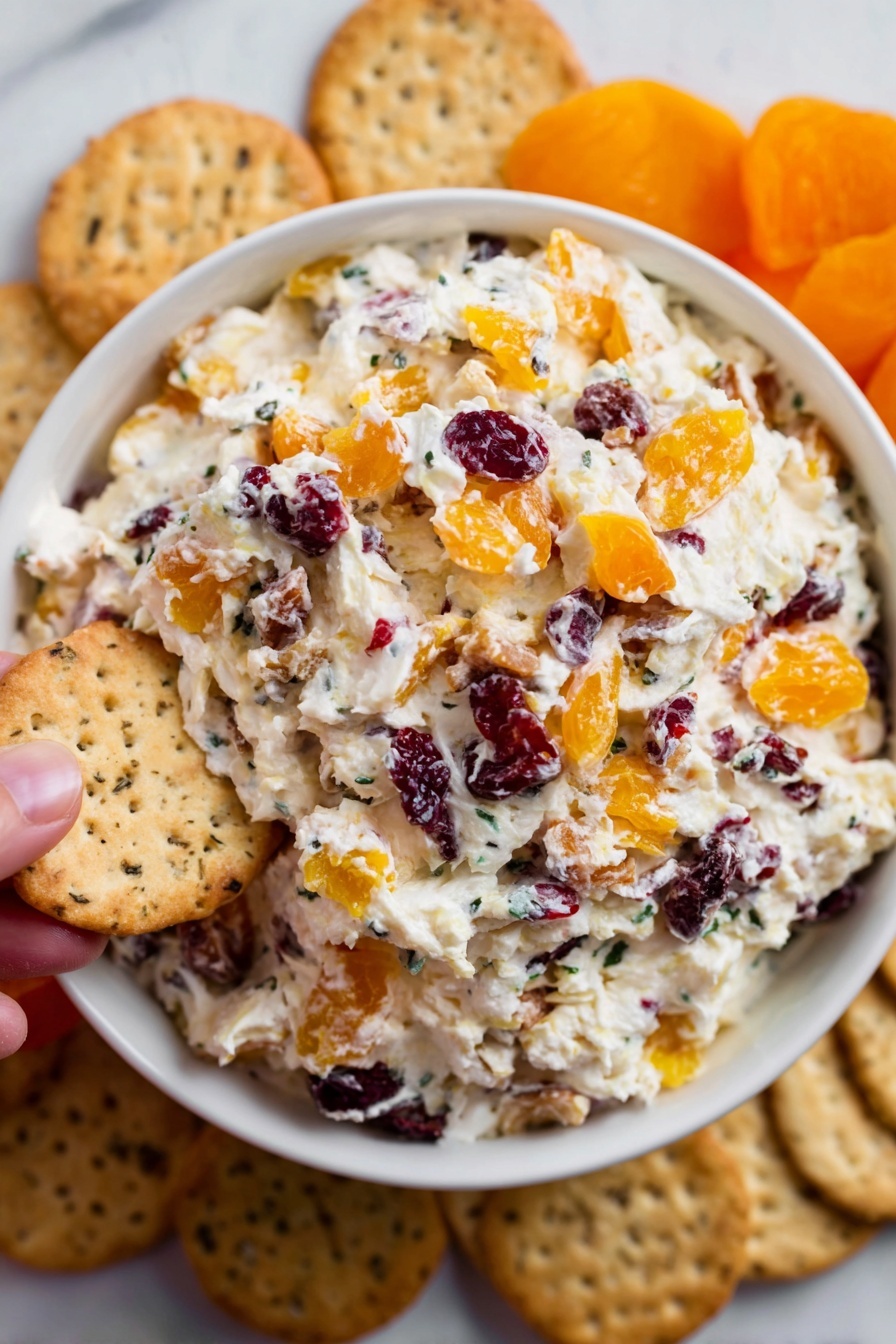 Cranberry Orange Cream Cheese Spread, cranberry orange dip, holiday cream cheese appetizer, quick fruit spread, festive cheese dip - A white bowl is filled with a creamy mixture that has three layers of visible ingredients: the base is soft white cream cheese, mixed throughout with chunks of orange dried apricots and small dark red dried cranberries, creating a colorful and textured look. Around the bowl, there are beige crackers placed on a white marbled surface, some showing a speckled texture. A woman's hand is holding one of the crackers near the edge of the bowl. The scene is bright and clear, showing the fresh, inviting look of the snack. photo taken with an iphone --ar 2:3 --v 7