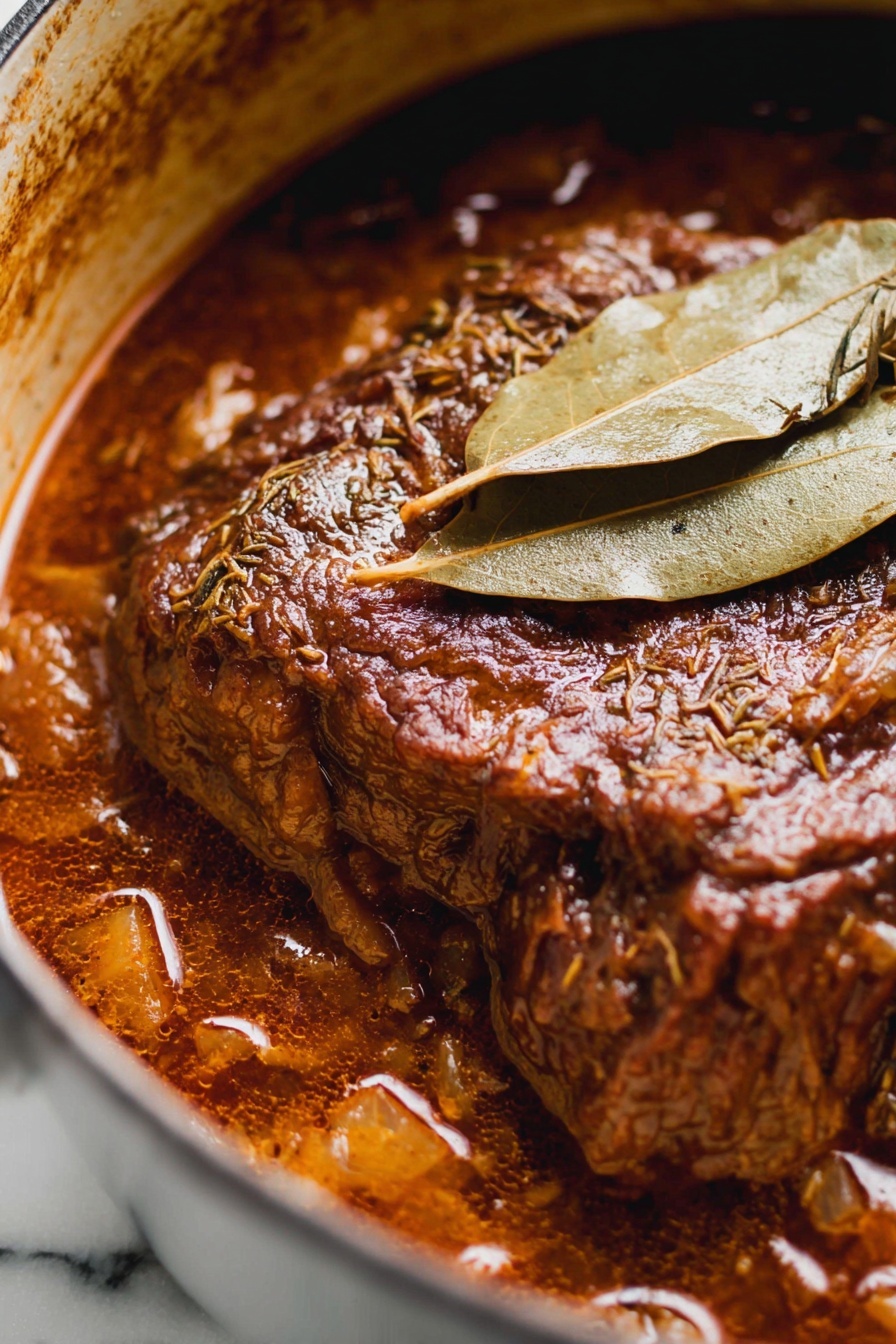 French Onion Chuck Roast, French Onion Beef Roast, slow cooker onion roast, caramelized onion beef, hearty beef roast recipe - The image shows a close-up of a rich, dark brown meat stew in a pot, with the main piece of meat at the center, covered in thick sauce. The meat has a textured surface with visible herbs and spices, and it looks tender and juicy. Two dry, light brown bay leaves rest on top of the meat, adding a layer of texture and color contrast. The stew sauce has a glossy finish with chunks visible around the edges, and the pot’s inner side is stained in a brownish color from the cooking. The background is a white marbled texture. photo taken with an iphone --ar 2:3 --v 7