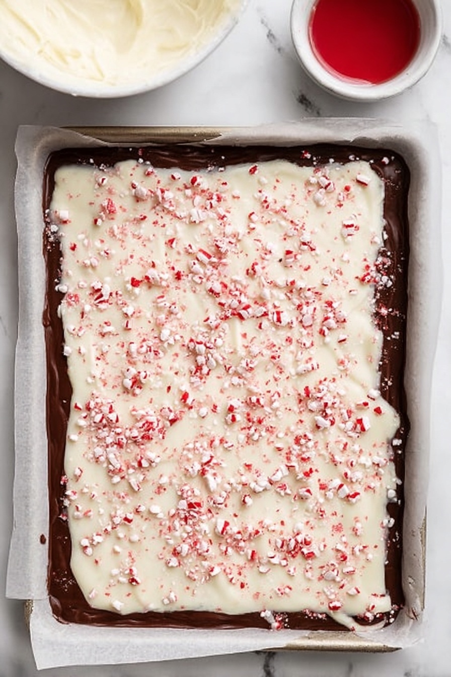 Peppermint Bark, Peppermint Bark Recipe, Holiday Chocolate Treats, Mint Chocolate Candy, Easy Festive Dessert - A rectangular baking tray lined with parchment paper holds a layered peppermint bark. The bottom layer is dark chocolate, smooth and shiny with slightly uneven edges extending close to the parchment's border. On top, there is a thick layer of white chocolate, creamy and textured, spread evenly but with natural ripples. The white chocolate layer is sprinkled generously with small, crushed red peppermint pieces scattered across the entire surface, adding a festive red and white contrast. In the top left corner of the image, a small white bowl with a creamy white mixture is partially visible, and a small white bowl with red liquid is nearby on a surface that looks like white marble. Photo taken with an iphone --ar 2:3 --v 7