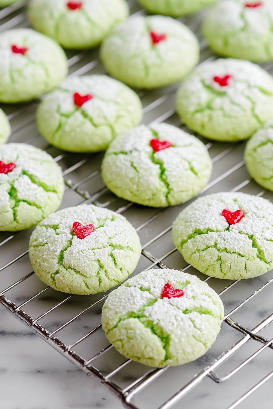 Green Peppermint Cake Mix Cookies, peppermint cookie recipe, festive holiday cookies, easy mint chocolate cookies, winter dessert ideas - The image shows rows of light green cookies on a silver cooling rack, each cookie round and cracked on top with a dusting of white powdered sugar. Each cookie has a small red heart-shaped decoration placed near the center. The cooling rack and cookies sit on a white marbled surface that is smooth and clean. The cookies appear soft and slightly puffy with a delicate texture. Photo taken with an iphone --ar 2:3 --v 7