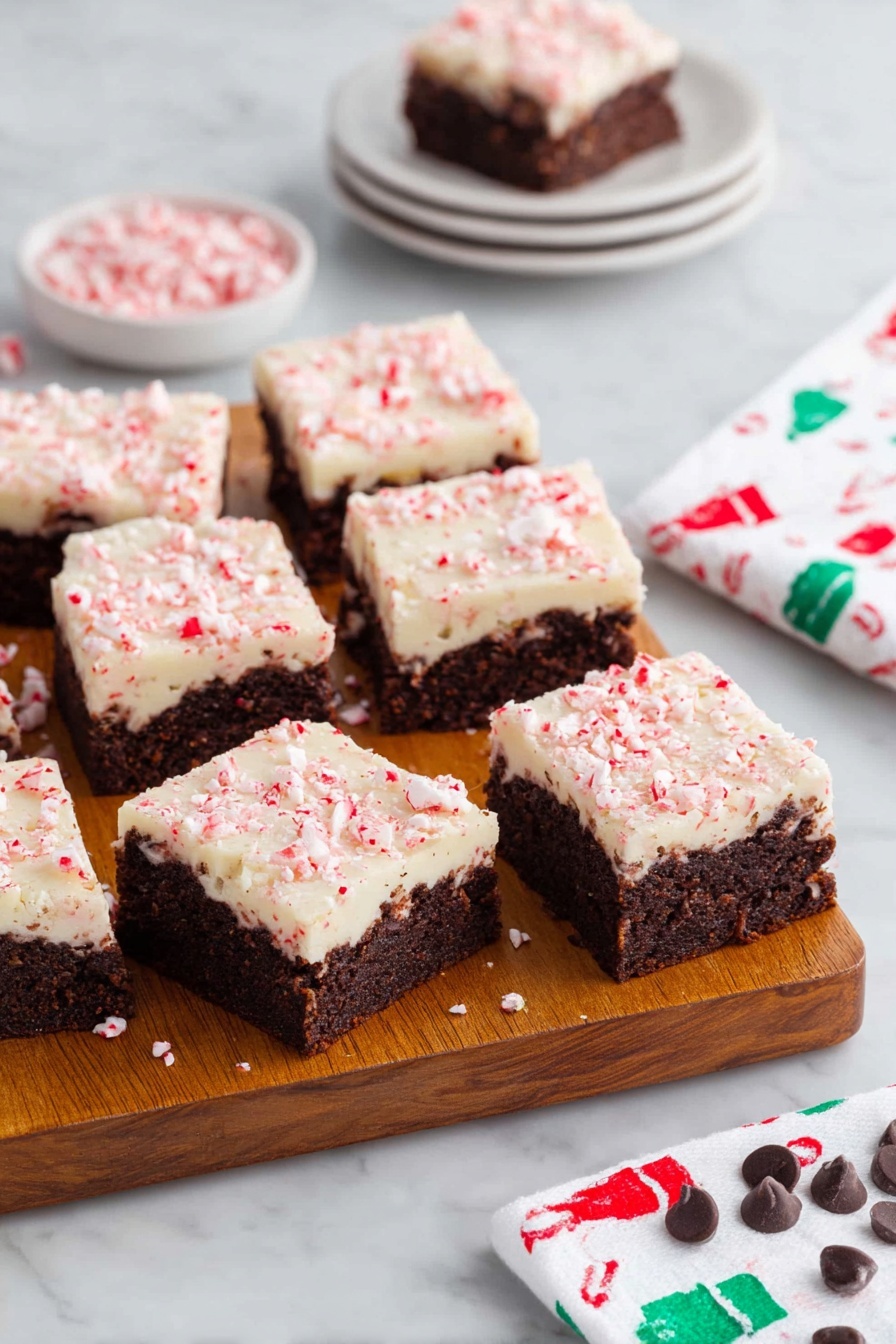 Peppermint Bark Brownies, Chocolate Mint Desserts, Holiday Brownie Recipes, Fudgy Mint Brownies, Festive Brownie Treats - A wooden board holds eleven square brownies, each with two layers: a dark brown, dense textured base with visible chunks inside, topped by a creamy white layer sprinkled with crushed pink peppermint pieces giving a slightly rough texture. Three brownies are separated at the front, showing clear edges and layers. Behind the board, there is a small white stack of four plates with one brownie on top, and to the right, a white napkin with a pattern of small green and red chef hats rests on a white marbled surface. Scattered peppermint candies and a few chocolate chips decorate the bottom edge of the surface. Photo taken with an iphone --ar 2:3 --v 7