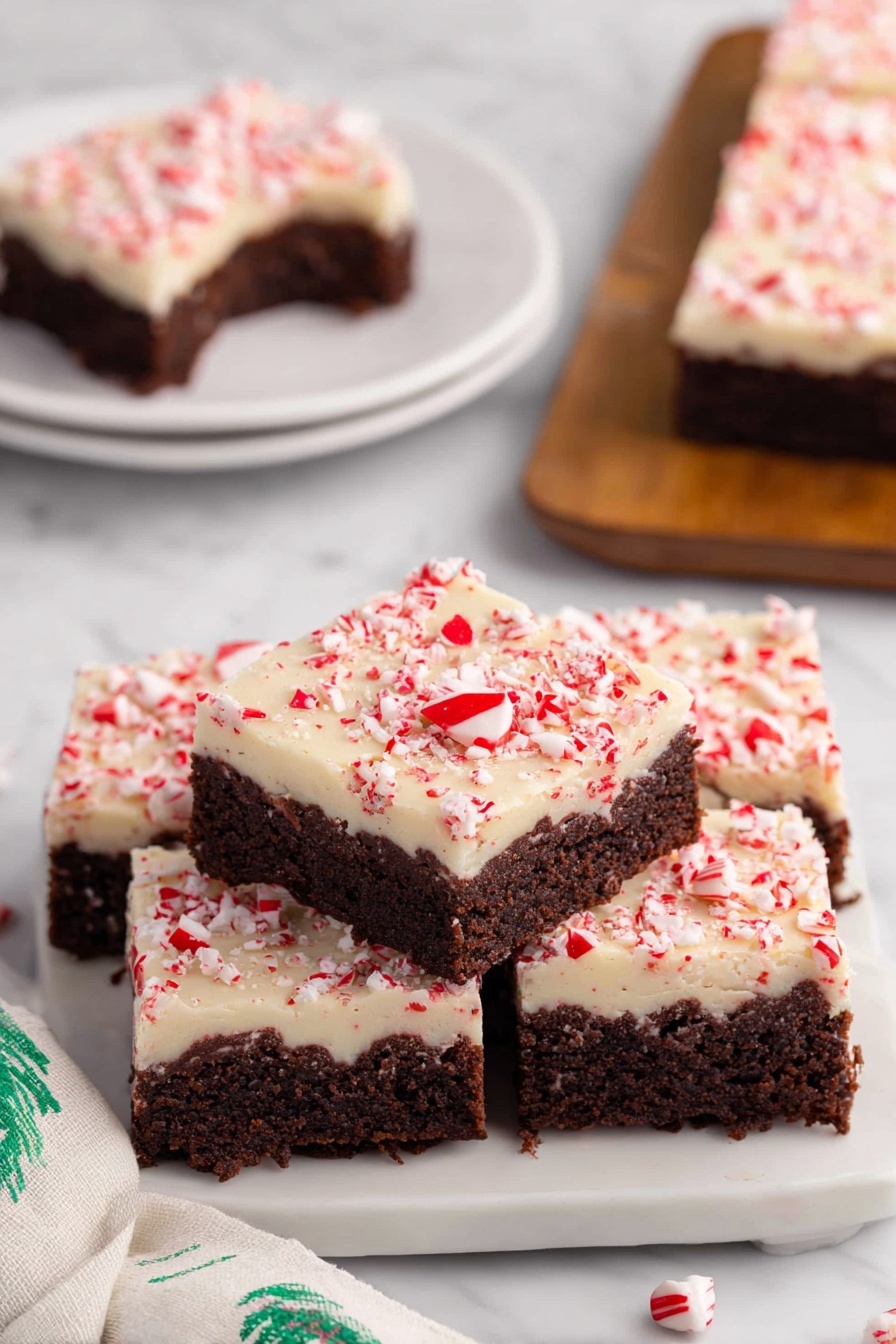 Peppermint Bark Brownies, Chocolate Mint Desserts, Holiday Brownie Recipes, Fudgy Mint Brownies, Festive Brownie Treats - The image shows a stack of four square peppermint brownies on a white plate with a white marbled surface underneath. Each brownie has two layers: a thick, dark brown chocolate base with a moist, dense texture, and a creamy white topping sprinkled with crushed red peppermint bits that add a pop of color and texture. One brownie on top has a small bite taken out, revealing the rich chocolate layer inside. In the background, there is a similar white plate with another brownie on it, and a soft white cloth with green patterns is visible on the side. photo taken with an iphone --ar 2:3 --v 7