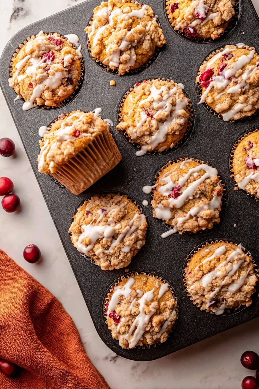 Cranberry Orange Muffins, easy cranberry orange muffin recipe, healthy cranberry muffin ideas, fall breakfast muffins, citrus cranberry baked goods - The image shows a dark muffin tin with nine muffins inside it, all golden brown with a crumbly topping that is light tan and unevenly spread over each muffin. Bright red berry pieces peek through the muffins, adding spots of color. White glaze is drizzled in thin lines over the top of each muffin, contrasting with the crumbly texture. One muffin is pulled slightly out of its spot showing the sides, which have a ridged paper liner of a light brown color. The muffin tin rests on a white marbled surface with a soft orange cloth partially visible in the bottom left corner and a few dark red whole berries scattered near the tin. photo taken with an iphone --ar 2:3 --v 7