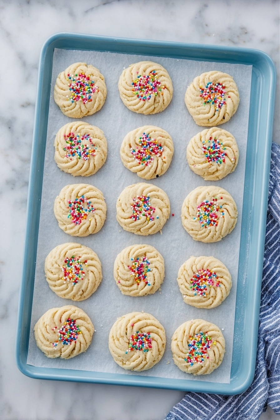 Butter Cookies, Easy Butter Cookies Recipe, Buttery Cookie Recipe, Classic Butter Cookies, Soft and Crispy Butter Cookies - There are fifteen round cookies placed on parchment paper on a light blue baking tray. The cookies have a swirled pattern with textured ridges in a pale beige color. Nine of the cookies are sprinkled with colorful round sprinkles, while six cookies have no sprinkles and show only the smooth beige dough. The tray is set on a white marbled surface with a blue and white striped cloth partially visible on the right side. photo taken with an iphone --ar 2:3 --v 7