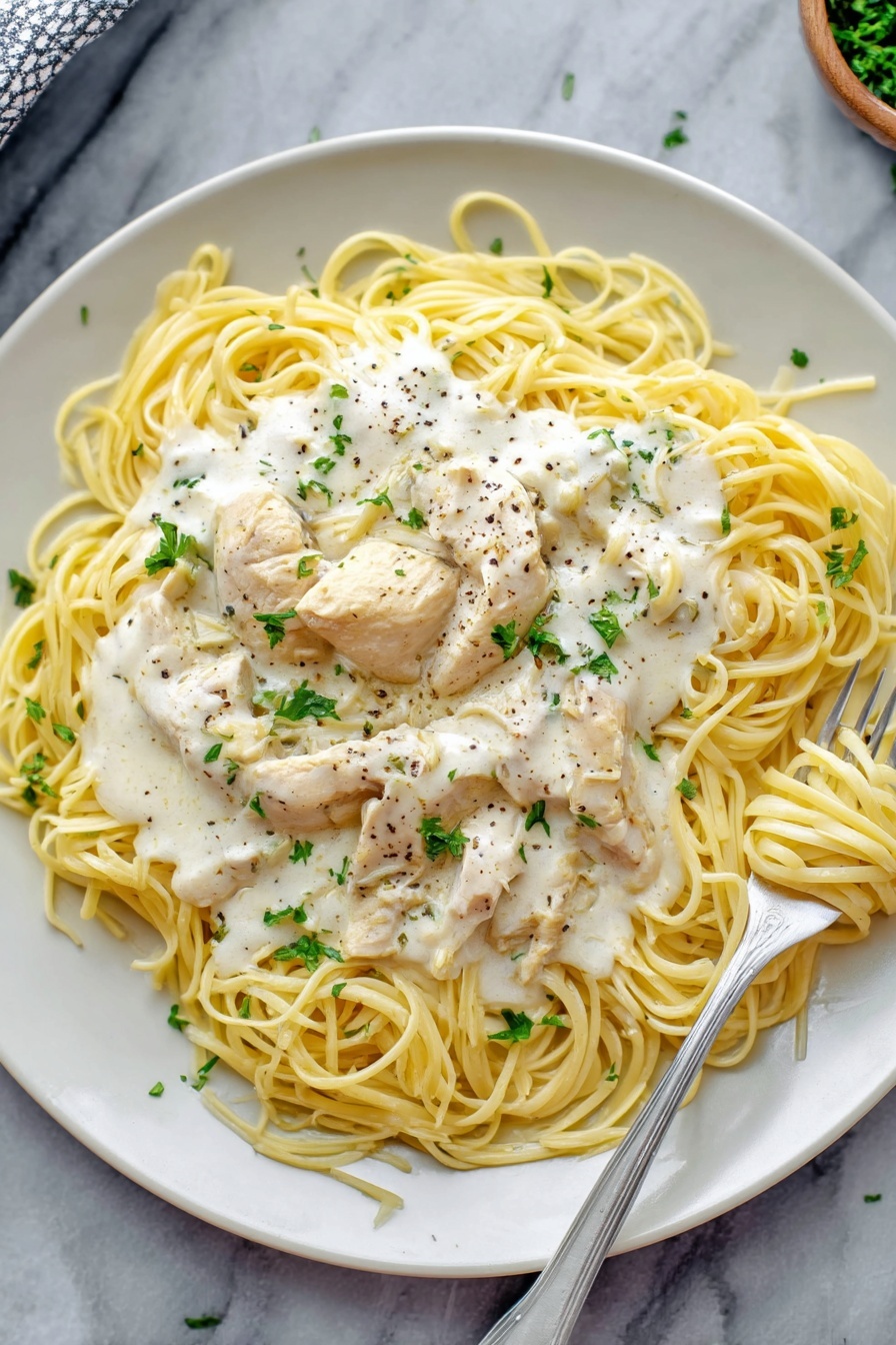 Creamy Crock Pot Angel Chicken, crock pot chicken recipes, easy slow cooker chicken, comforting chicken pasta dish, simple crockpot dinner - A white plate holds a dish of thin yellow spaghetti noodles arranged loosely in a circle. On top of the noodles are several pieces of light cream chicken, covered with a thick white creamy sauce that has small bits of herbs and black pepper sprinkled over it. Fresh green parsley leaves are scattered on top of the sauce for a touch of color. A silver fork rests on the right side of the plate, twirling some noodles with sauce. The plate sits on a white marbled surface. Photo taken with an iphone --ar 2:3 --v 7