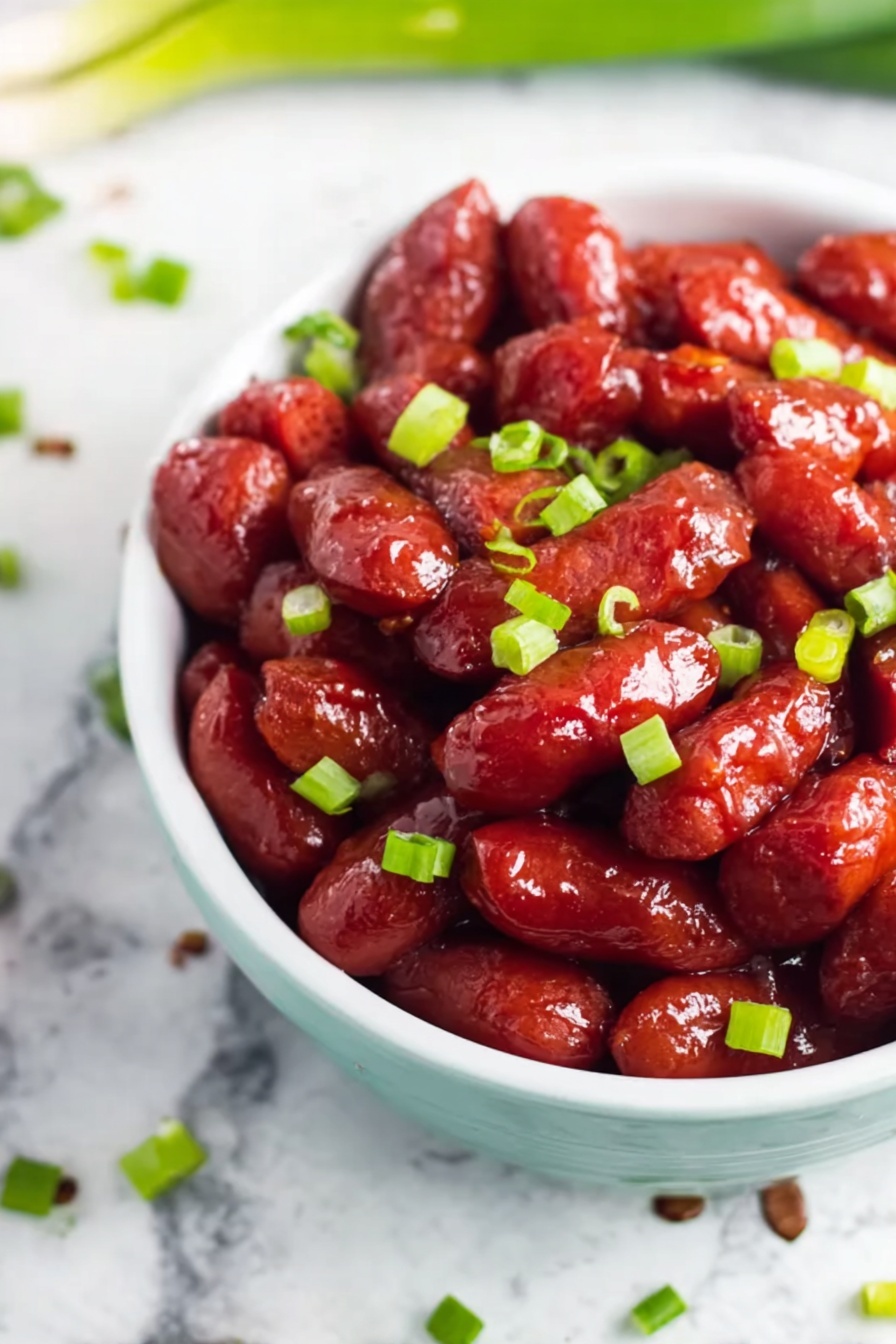 Slow Cooker BBQ Little Smokies, BBQ Little Smokies recipe, easy slow cooker snacks, game day appetizer ideas, smokie sausage appetizer - The image shows a white bowl filled with small, shiny red sausage-like pieces covered in a glossy sauce, giving them a sticky and rich appearance. Bright green chopped scallions are scattered evenly over the sausages, adding a fresh contrast in color. The bowl sits on a white marbled surface with a few scattered scallions around it, creating a clean and simple background. The focus is close up and clear, highlighting the texture and color of the sausages and scallions. photo taken with an iphone --ar 2:3 --v 7