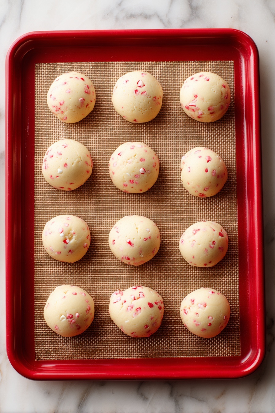 White Chocolate Candy Cane Cookies, holiday cookie recipes, mint chocolate cookies, Christmas cookie ideas, festive dessert recipes - The image shows eight round cookie dough balls placed on a brown baking mat lined inside a red-edged tray. Each cookie ball has a creamy, pale dough base with small red specks all over, and some white chocolate chips peek through the dough, adding texture. The surface under the baking mat is a white marbled texture. The dough looks soft, and the balls are evenly spaced apart. photo taken with an iphone --ar 2:3 --v 7