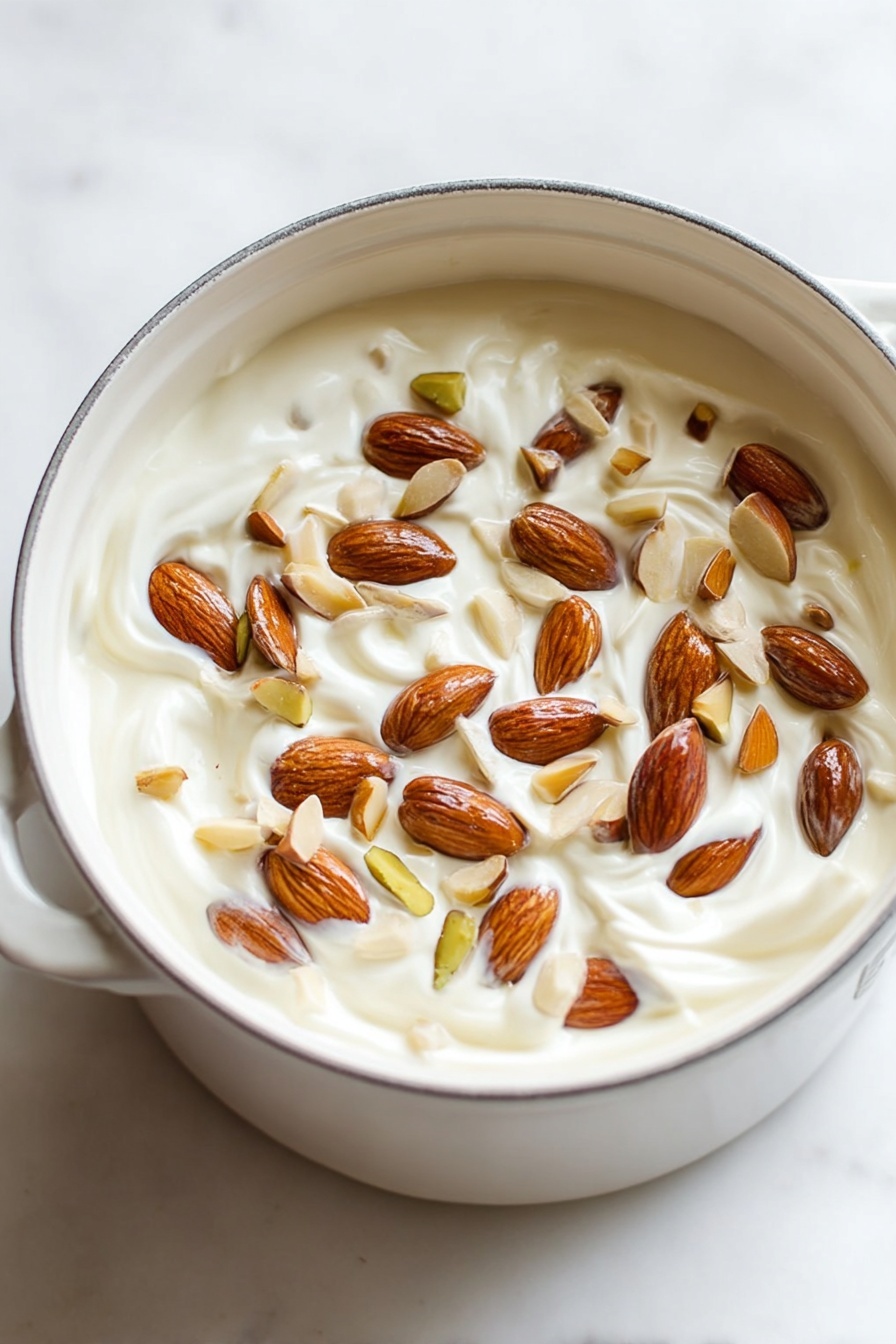 Italian Nut Torrone, Nut Torrone recipe, Italian nougat with nuts, homemade torrone, traditional Italian sweets - A white pot filled with one main layer of thick, creamy white yogurt mixed with whole and sliced almonds scattered evenly on the top. The almonds vary in shades of light brown to dark brown and are embedded slightly into the creamy yogurt, creating a textured surface with visible swirls in the yogurt beneath. The pot is sitting on a white marbled surface with soft lighting that highlights the smoothness of the yogurt and the natural shine of the nuts. Photo taken with an iphone --ar 2:3 --v 7