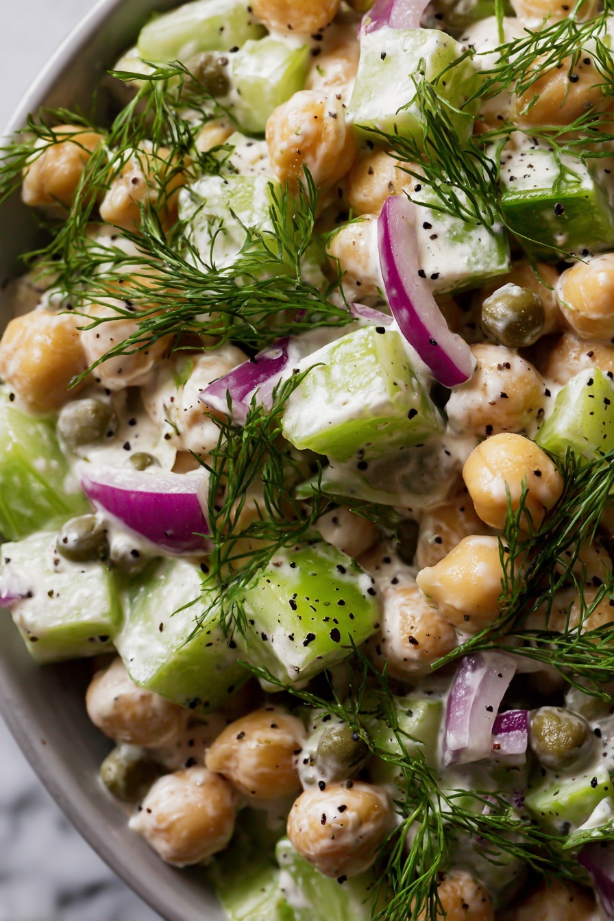 Healthy Tuna Chickpea Salad, Tuna Chickpea Salad, Healthy Lunch Recipes, Quick Healthy Salads, nutritious tuna salad - The image shows a close-up of a salad with several layers and colors. The main elements are round chickpeas that are beige and smooth, mixed with small cubes of light green celery and purplish red onion. There are round, green capers scattered throughout. Fresh green dill sprigs are placed on top, adding a feathery texture. The salad is coated with a creamy, white dressing that has small black seeds mixed in. The entire dish sits against a white marbled surface in soft light. photo taken with an iphone --ar 2:3 --v 7