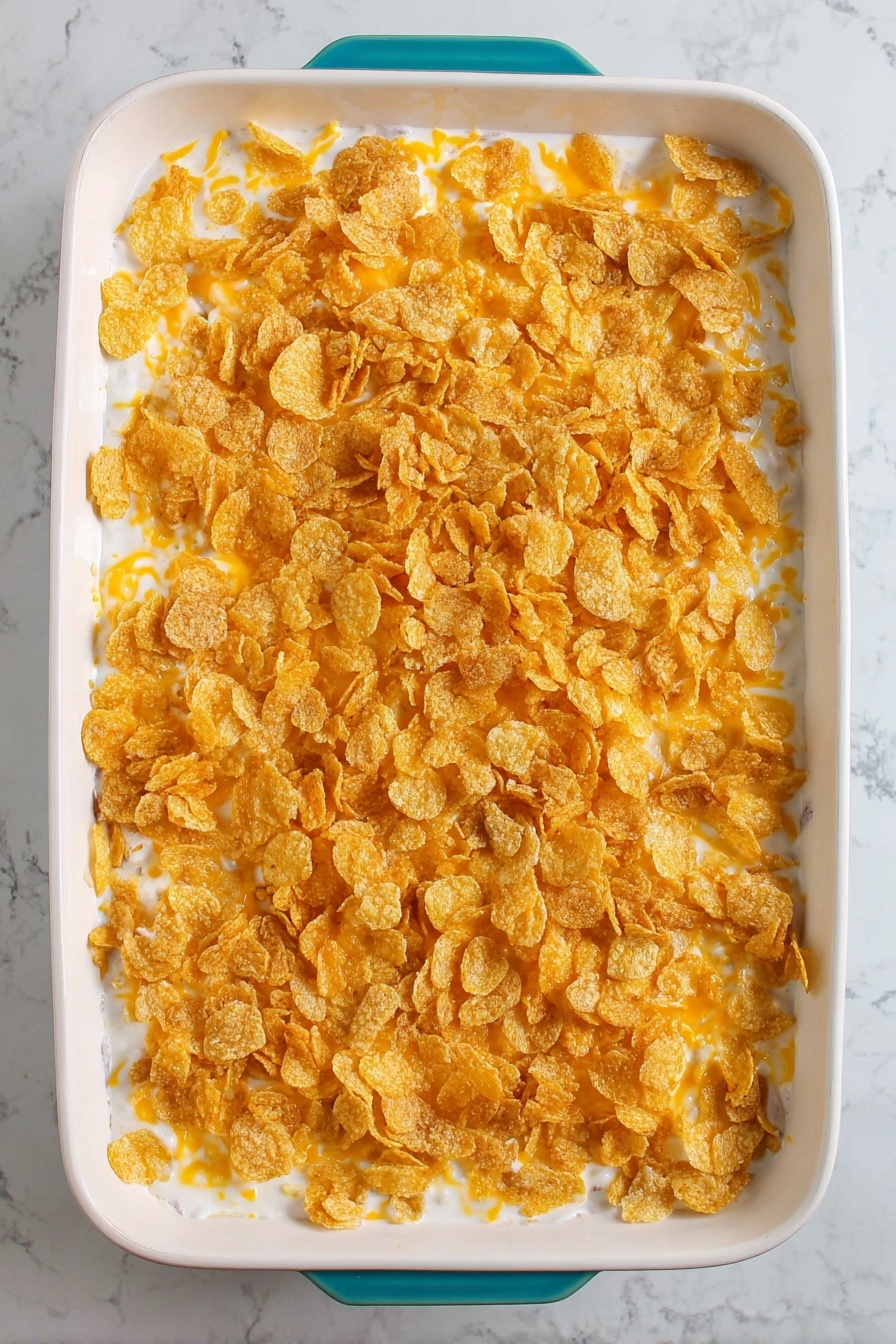 Cheesy Hashbrown Casserole, cheesy potato casserole, baked hashbrown recipe, cheesy potato bake, comfort food casserole - The image shows a white rectangular casserole dish filled with a three-layered casserole. The bottom layer is a creamy white sauce with some small visible pieces of ingredients. The middle layer is light orange shredded cheese scattered evenly. The top layer is a generous covering of crunchy golden corn flakes spread all over. The dish is placed on a white marbled surface, and the dish has a teal handle barely visible at the bottom edge. Photo taken with an iphone --ar 2:3 --v 7