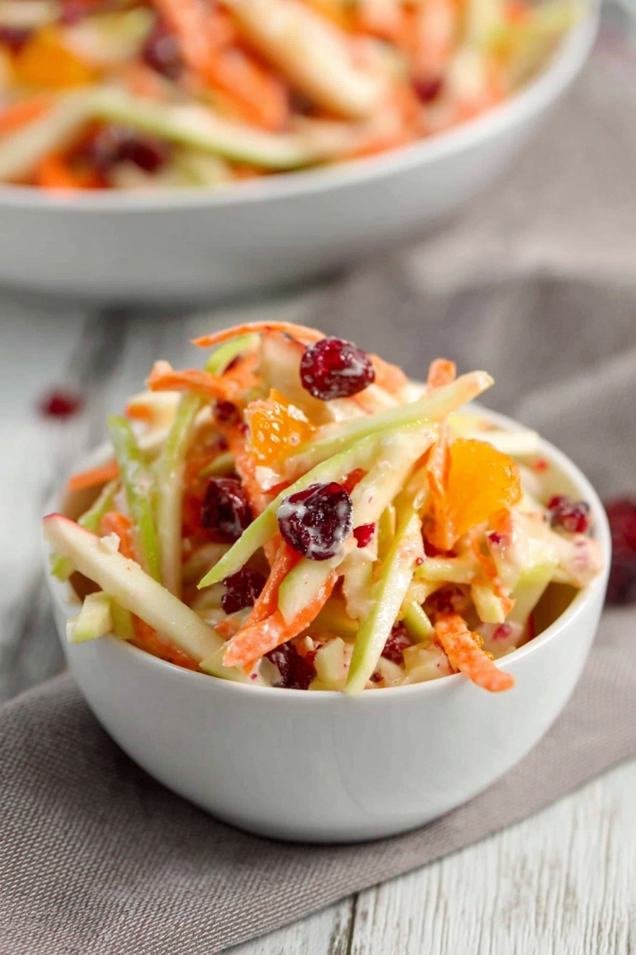 Carrot Apple Salad with Cranberries and Mandarin, healthy fruit salad with cranberries and mandarins, easy colorful vegetable salad, fresh apple and carrot side dish, quick fruit salad recipe - A clear glass bowl filled with a colorful salad sits on top of a wooden salad spoon and fork resting on a gray patterned cloth, all placed on a white marbled surface. The salad shows layers of thin orange carrot strips, creamy white slivers of apple, green apple slices, bright orange mandarin pieces, and dark red dried cranberries or similar fruit mixed throughout. Two small white bowls in front also hold the salad, showing the same colorful mixture with vibrant orange, green, red, and creamy white pieces clearly visible. photo taken with an iphone --ar 2:3 --v 7