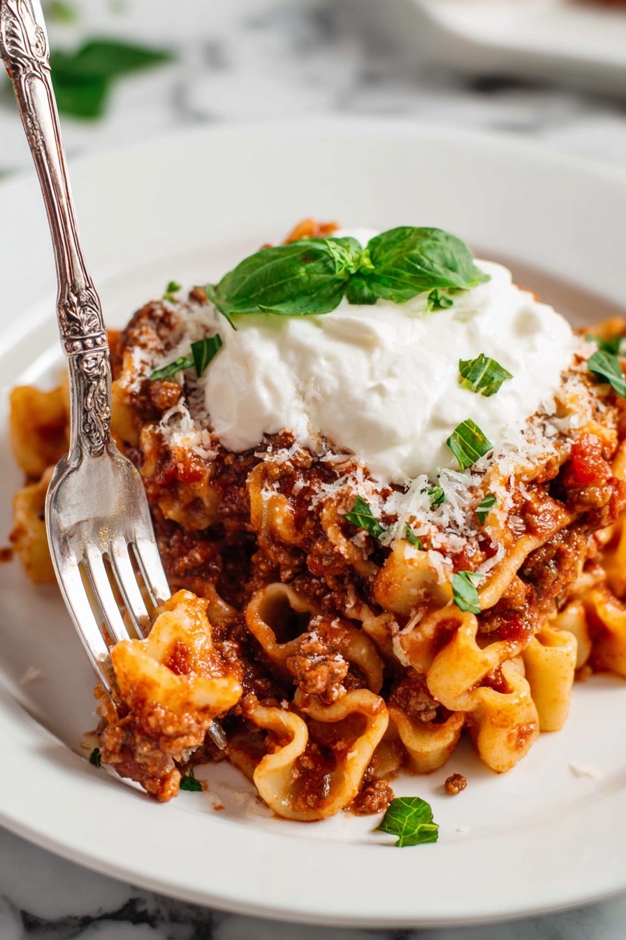 Easy Homemade Lasagna Hamburger Helper, creamy lasagna skillet meal, quick lasagna dinner, cheesy hamburger casserole, one-pot lasagna recipe - In the image, there is a white plate on a white marbled surface with pasta mixed with a thick red-brown meat sauce spread across it. On top of the pasta, there is a dollop of white cream with a small green basil leaf placed on it. Thin green basil strips and a sprinkle of shredded white cheese are scattered around the pasta on the plate. A silver fork rests on the edge of the plate. Behind this plate, a larger white bowl filled with more of the pasta dish sits on a light cloth, garnished with a whole basil leaf in the center. To the upper right, part of another white plate with the same pasta dish is visible. Small red cherry tomatoes are scattered at the bottom right corner on the white marbled surface, and a small shaker with red pepper flakes is placed to the left of the main plate. The scene is cozy and inviting, all set on the clean white marble background. photo taken with an iphone --ar 2:3 --v 7