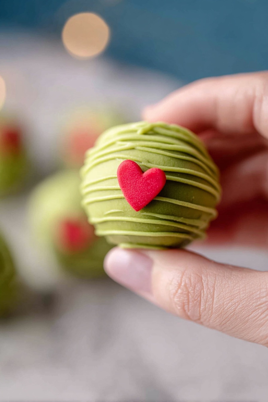Grinch Oreo Balls with Red Velvet Cookies, festive holiday treats, Christmas dessert, easy holiday truffles, Grinch-themed candies - This image shows a white bowl filled with round green balls that have smooth surfaces and are drizzled with thin lines of a lighter green shade. Each ball is topped with a small red heart-shaped decoration. The bowl rests on a matching white plate, both placed on a white marbled surface. Around the plate are a few more of the red heart shapes scattered softly. The background is softly blurred with soft warm lights and out-of-focus colorful holiday decorations visible. Photo taken with an iphone --ar 2:3 --v 7