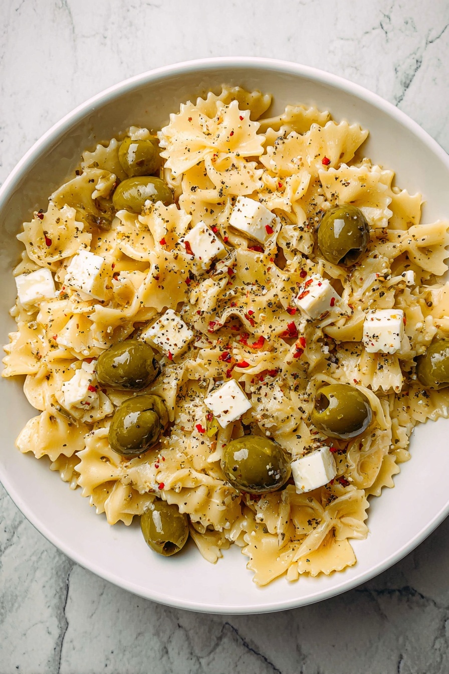 Dirty Martini Pasta Salad, pasta salad with olive brine, gourmet pasta salad, easy cocktail-inspired pasta dish, savory pasta salad recipes - A white bowl filled with pasta made of small, square-shaped pieces with wavy edges, light yellow in color, creating the base layer. Mixed evenly on top are green olives with small pieces of red inside them and small white cubes of cheese scattered throughout. The whole dish is sprinkled with black pepper, adding small dark specks. The bowl is placed on a white marbled surface. photo taken with an iphone --ar 2:3 --v 7