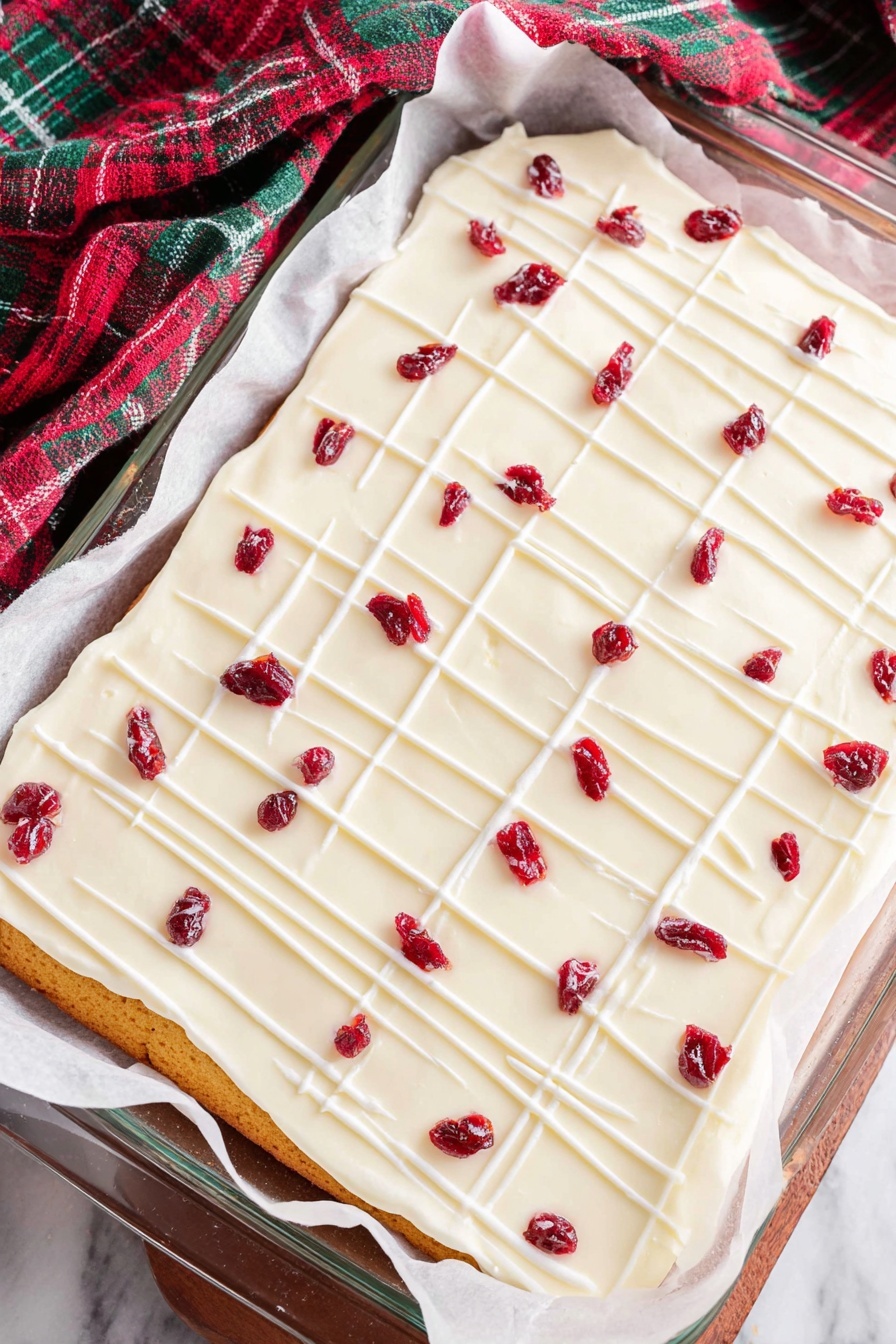 Cranberry White Chocolate Bliss Bars, festive dessert recipes, easy holiday treats, cranberry and white chocolate bars, no-bake holiday desserts - A rectangular layered dessert lies on white parchment paper inside a glass baking dish on a white marbled surface. The bottom layer is golden brown, thin, and even. On top is a thick, smooth white frosting layer covering the entire surface. Thin white icing lines are drizzled across the frosting, forming a light grid pattern. Scattered over the top are small dried red fruit pieces, evenly spaced, adding bright red spots to the white frosting. The cloth nearby has green, red, and dark colors in a check pattern. photo taken with an iphone --ar 2:3 --v 7