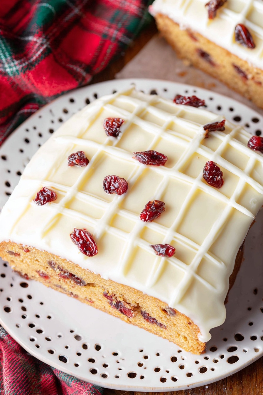 Cranberry White Chocolate Bliss Bars, festive dessert recipes, easy holiday treats, cranberry and white chocolate bars, no-bake holiday desserts - A single slice of cake is shown on a white decorative plate with small cut-out patterns. The cake has two clear layers: a dense golden-brown base with visible small dark red dried fruit pieces inside, and a thick smooth white icing layer on top, decorated with a white grid pattern and scattered dark red dried fruit pieces. The plate rests on a wooden surface with a blurred part of the whole cake and a red and green fabric in the background. Photo taken with an iphone --ar 2:3 --v 7