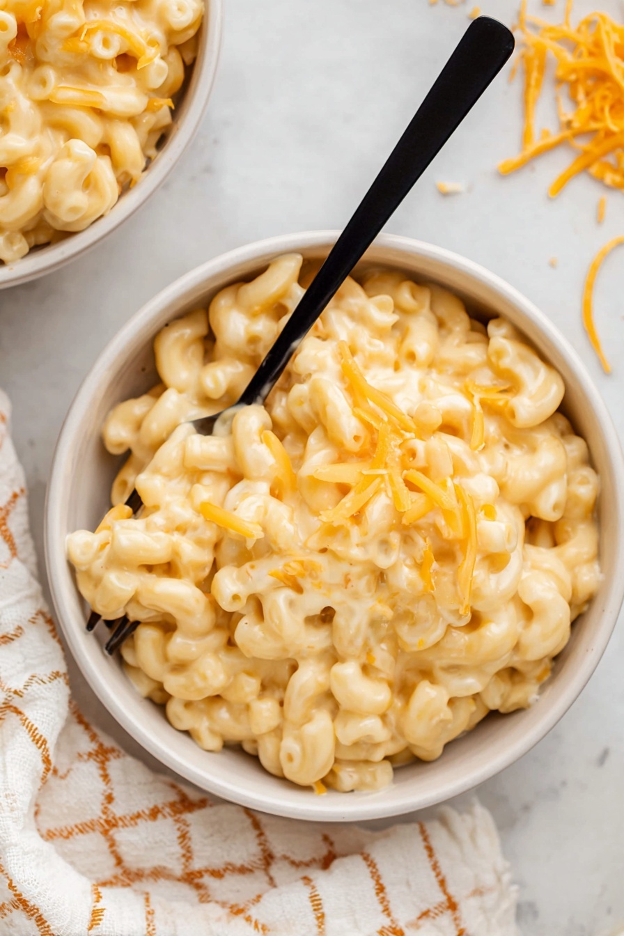 Stovetop Mac and Cheese, creamy mac and cheese, cheesy pasta recipe, one-pot mac and cheese, homemade mac and cheese - A close-up view of creamy macaroni and cheese inside a white pot with a black rim. The macaroni is elbow-shaped, covered in smooth, pale yellow cheese sauce, with some small melted orange cheese bits sprinkled on top. A wooden spoon with a visible grain is scooping a portion of the cheesy macaroni, lifting it slightly above the pot. The pot sits on a light fabric with a yellow and white geometric pattern, all on a white marbled surface. Photo taken with an iphone --ar 2:3 --v 7