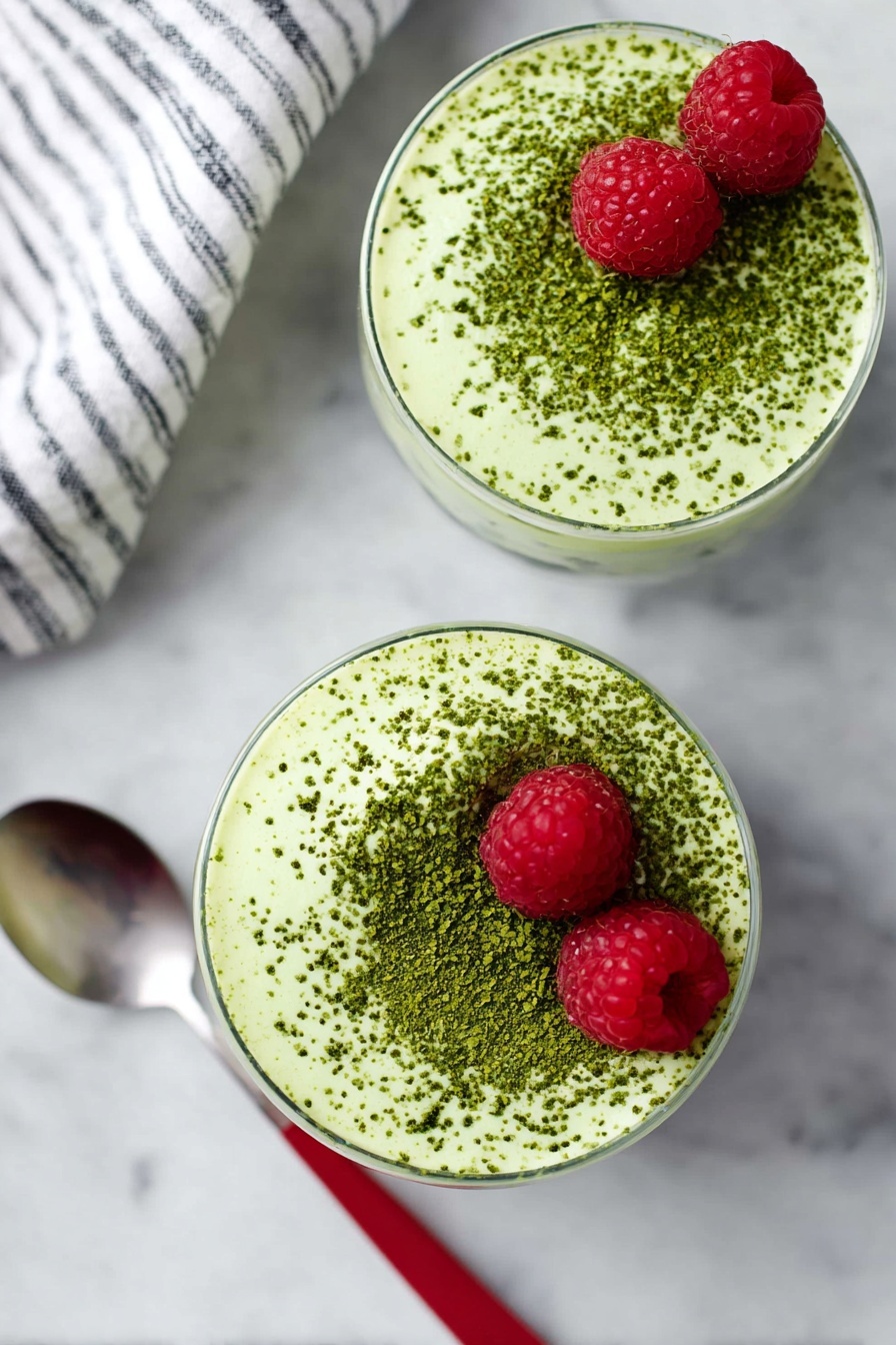 Matcha Chia Protein Pudding, healthy matcha pudding, vegan protein breakfast, quick nutritious snack, plant-based chia pudding - Two round glasses filled with a creamy light green mousse topped with a dusting of darker green powder evenly spread on the surface. Each mousse has two bright red raspberries placed near the top edge. The glasses sit on a white marbled surface. A spoon with a red handle is placed next to the bottom glass. Part of a white and black striped cloth is visible on the top left corner. Photo taken with an iphone --ar 2:3 --v 7