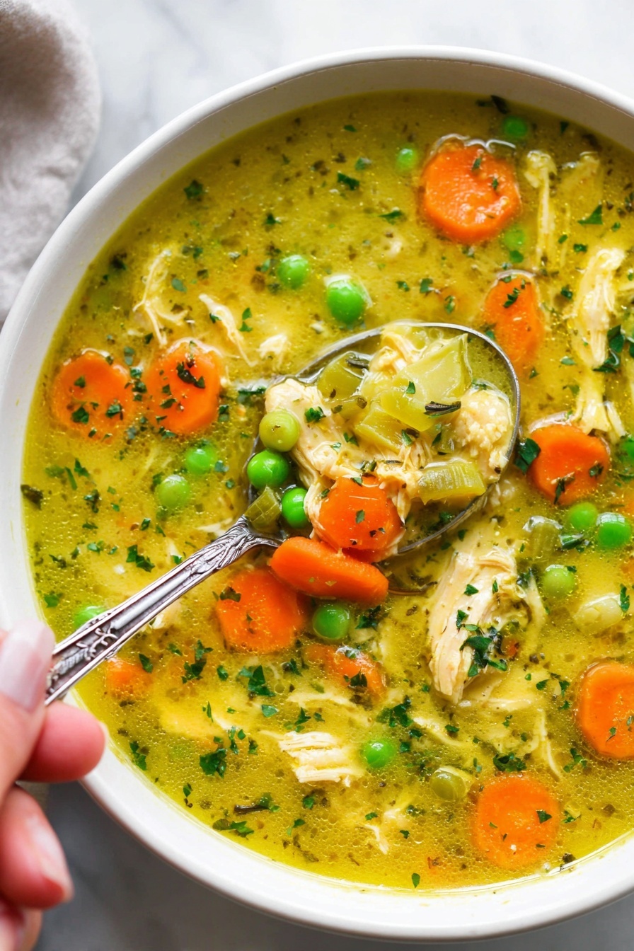 Turmeric Chicken Soup with Coconut Milk, turmeric chicken soup, coconut milk soup recipes, healthy chicken soup, cozy comfort soups - The image shows a white bowl filled with a colorful chicken soup. The soup has a yellowish broth with small pieces of shredded chicken scattered throughout. Bright orange carrot slices and vibrant green peas float on the surface along with translucent chunks of celery. Small green herb bits are sprinkled all over, adding texture and color. A silver spoon with detailing on the handle is partially submerged in the soup, held by a woman's hand, scooping some of the ingredients. The bowl sits on a white marbled surface, giving a clean and fresh look. photo taken with an iphone --ar 2:3 --v 7