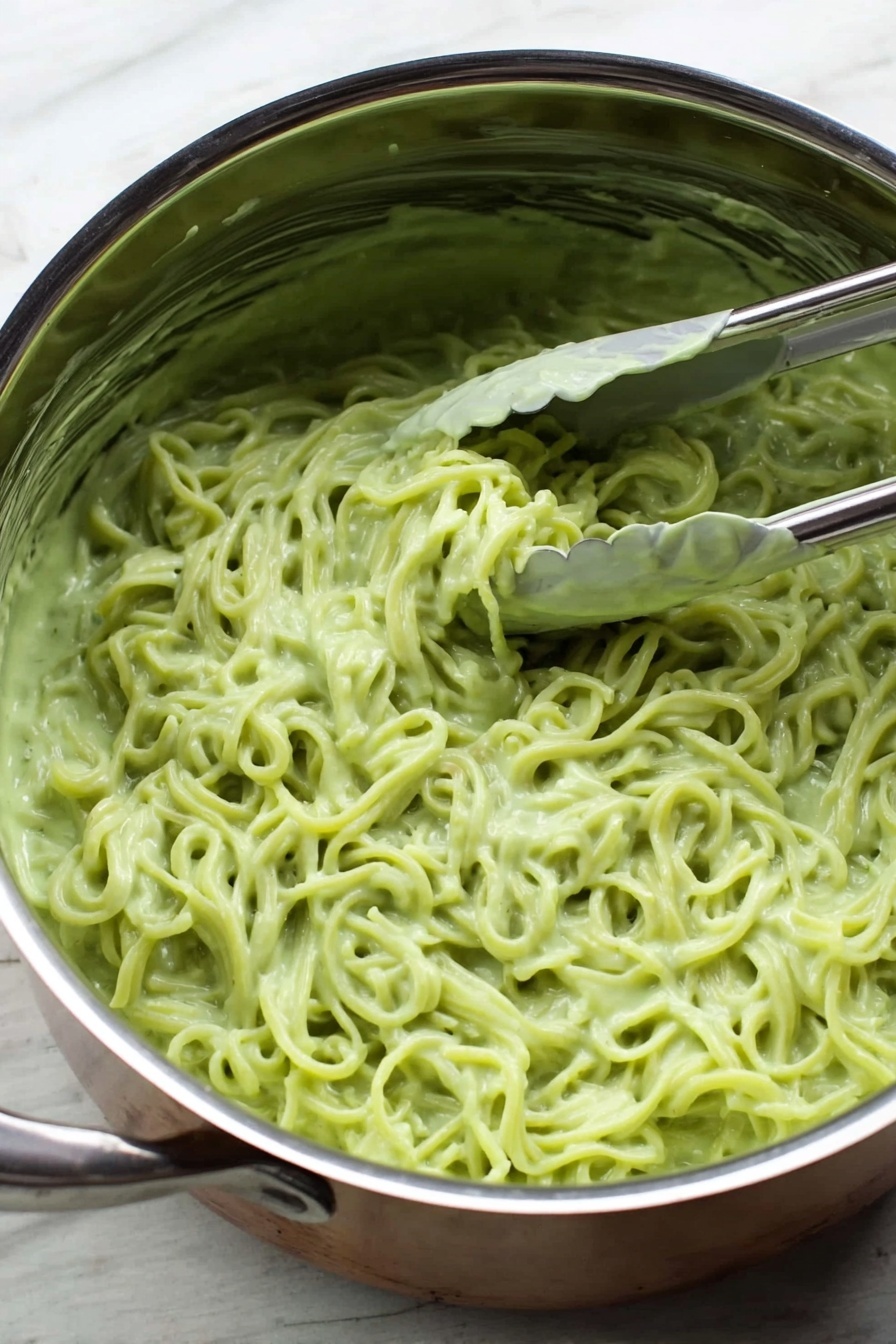 Vegan Cilantro Lime Noodles, vegan noodle recipes, healthy pasta dishes, quick plant-based meals, flavorful vegan noodle ideas - Inside a shiny silver pot placed on a white marbled surface, there is a mass of noodles covered in a thick, light green sauce. The sauce is creamy and clings to the noodles, giving them a smooth texture. A silver tong is partially dipped into the noodles on the right side of the pot, with a bit of sauce stuck on the tong. The noodles have a slightly twisted shape and fill most of the pot inside. Photo taken with an iphone --ar 2:3 --v 7