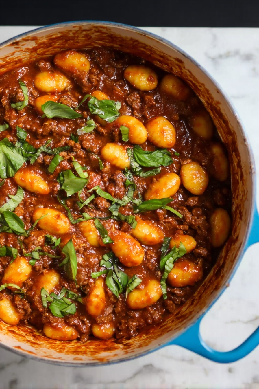 Gnocchi Bolognese, homemade gnocchi with Bolognese sauce, easy Italian gnocchi recipe, comforting Bolognese pasta, hearty gnocchi dish - The image shows a blue pot filled with a rich brown meat sauce mixed with soft, yellow-orange gnocchi. The top layer is scattered with pieces of fresh green herbs, adding a bright contrast to the warm colors of the sauce and gnocchi. The pot's handle is visible on the right side, resting on a white marbled surface. The texture of the gnocchi looks smooth and tender, while the meat sauce appears thick and hearty, covering all pieces evenly. Photo taken with an iphone --ar 2:3 --v 7