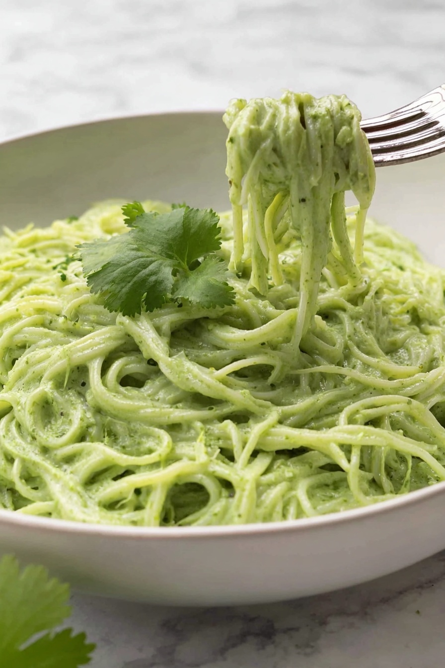 Vegan Cilantro Lime Noodles, vegan noodle recipes, healthy pasta dishes, quick plant-based meals, flavorful vegan noodle ideas - This image shows a white bowl filled with creamy green noodles, with about two layers of noodles visible, all coated evenly in a thick, smooth green sauce. On top of the noodles, there is a single thin slice of lime placed near the center, with bits of black pepper sprinkled across the noodles. Next to the lime slice, a small bunch of fresh green cilantro leaves rests gently. A silver fork is placed on the right edge of the bowl, partly inserted into the noodles. The bowl sits on a white marbled surface. Photo taken with an iphone --ar 2:3 --v 7
