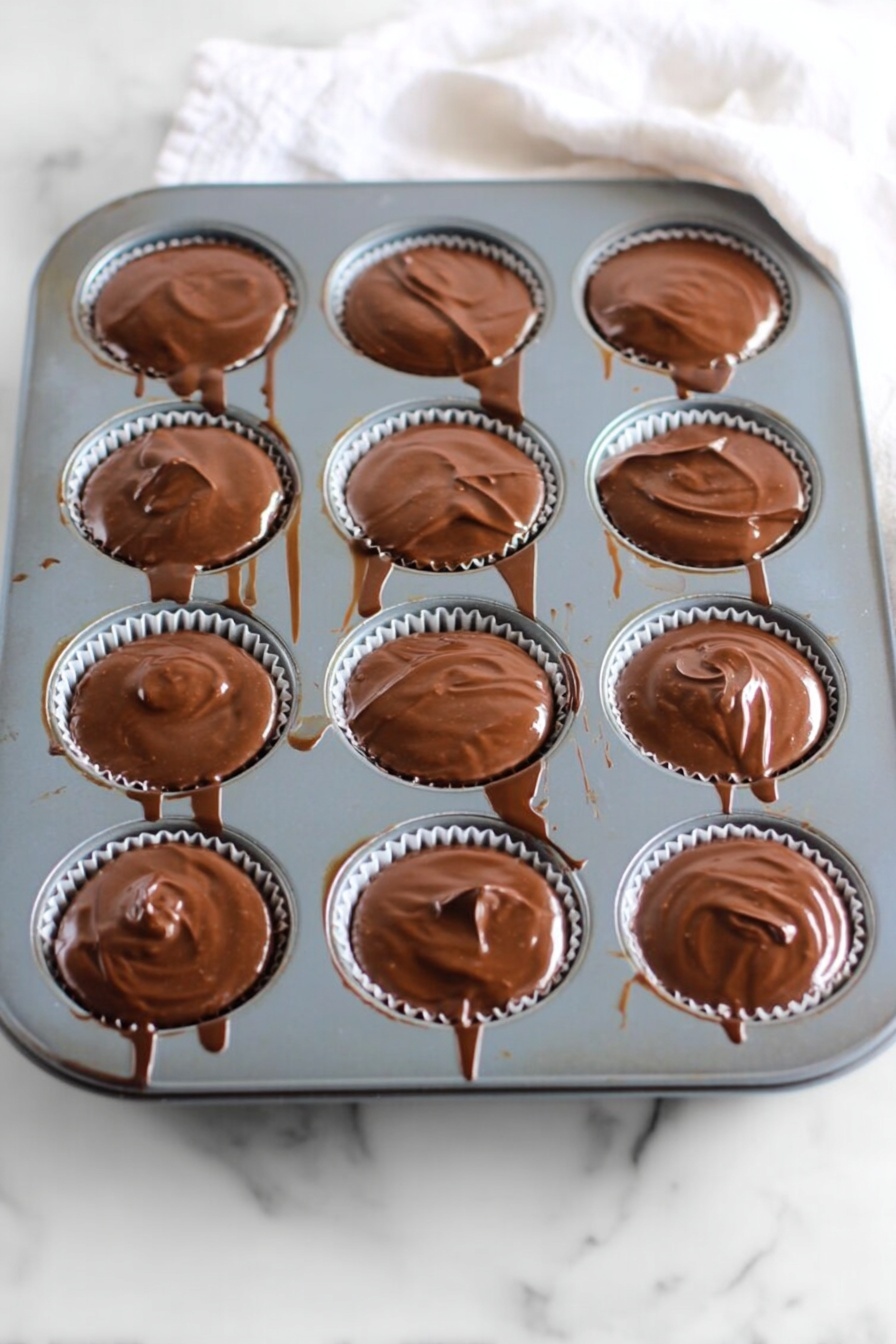 No Bake Peanut Butter Oat Cups, no bake oat snack, healthy peanut butter treats, simple no bake desserts, homemade peanut butter cups - A gray muffin tray with twelve round cups, each filled with a smooth, glossy layer of melted chocolate that slightly spills over the edges. The chocolate has a shiny, swirled texture on top, and thin chocolate drips and streaks connect some of the cups and run down the tray’s sides. The tray rests on a white marbled surface with a soft white cloth partially visible at the top right corner. Photo taken with an iphone --ar 2:3 --v 7