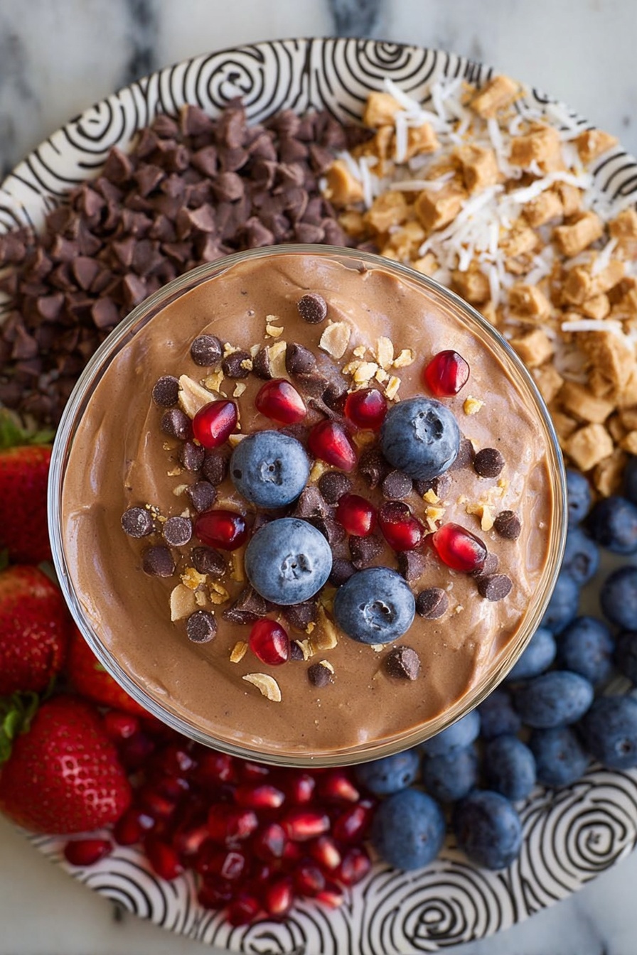 Chocolate Protein Fluff, healthy chocolate dessert, protein-packed snack, quick healthy treat, chocolate Greek yogurt dessert - A large round white plate with black spiral and zigzag patterns holds eight sections of colorful food arranged around a central glass bowl filled with smooth light brown chocolate mousse. Starting from the top left going clockwise, there are bright red whole strawberries with green tops, small dark brown chocolate chips, off-white shredded coconut, chopped pieces of chocolate peanut butter cups, bright red pomegranate seeds, dark blue blueberries, beige crunchy granola clusters, and then back to the strawberries. The plate is placed on a white marbled surface. Photo taken with an iphone --ar 2:3 --v 7