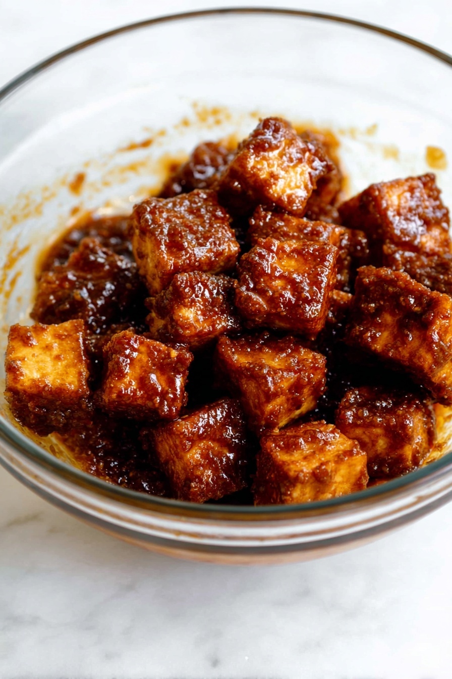 Crispy Gochujang Korean Tofu, Korean tofu stir-fry, spicy tofu recipe, vegan Korean dishes, crispy baked tofu - The image shows a clear glass bowl filled with several small cubes of fried tofu. Each tofu piece is coated in a dark brown, shiny sauce with a textured surface that looks sticky and flavorful. The tofu pieces are piled together in the bowl, some slightly stacked on top of others. The smooth surface of the glass bowl lets you see the sauce clinging to the sides. The bowl is placed on a white marbled surface. photo taken with an iphone --ar 2:3 --v 7