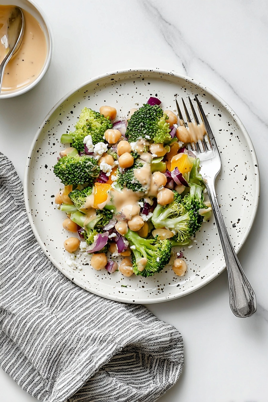 Chickpea Broccoli Salad with Lemon Tahini, healthy chickpea salad, vegan broccoli salad, lemon tahini dressing, quick vegetarian side dish - A white bowl filled with a colorful chopped salad showing three clear layers: a base layer of small broccoli florets and chunks of celery creating a dense green texture, a middle layer with light-colored chickpeas and bright orange bell pepper pieces scattered evenly, and a top layer with small cubes of purple onion and white crumbled cheese spread across, topped lightly with thin almond slices and a creamy tan dressing drizzled over. Around the bowl are a gray and white striped cloth, a white marbled surface, a spoon in a small jar of creamy dressing, a small gray dish with salt and pepper, and a fork and knife nearby, photo taken with an iphone --ar 2:3 --v 7